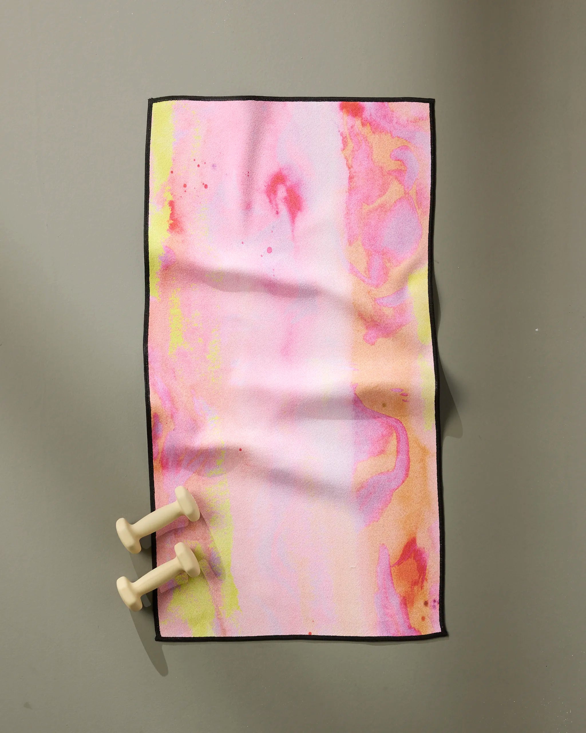 The GEOMETRY Purity towel, ultra-absorbent and fast-drying with pastel pink, yellow, and orange abstract patterns, lies flat on a gray surface. Two small beige dumbbells rest in the bottom left corner.