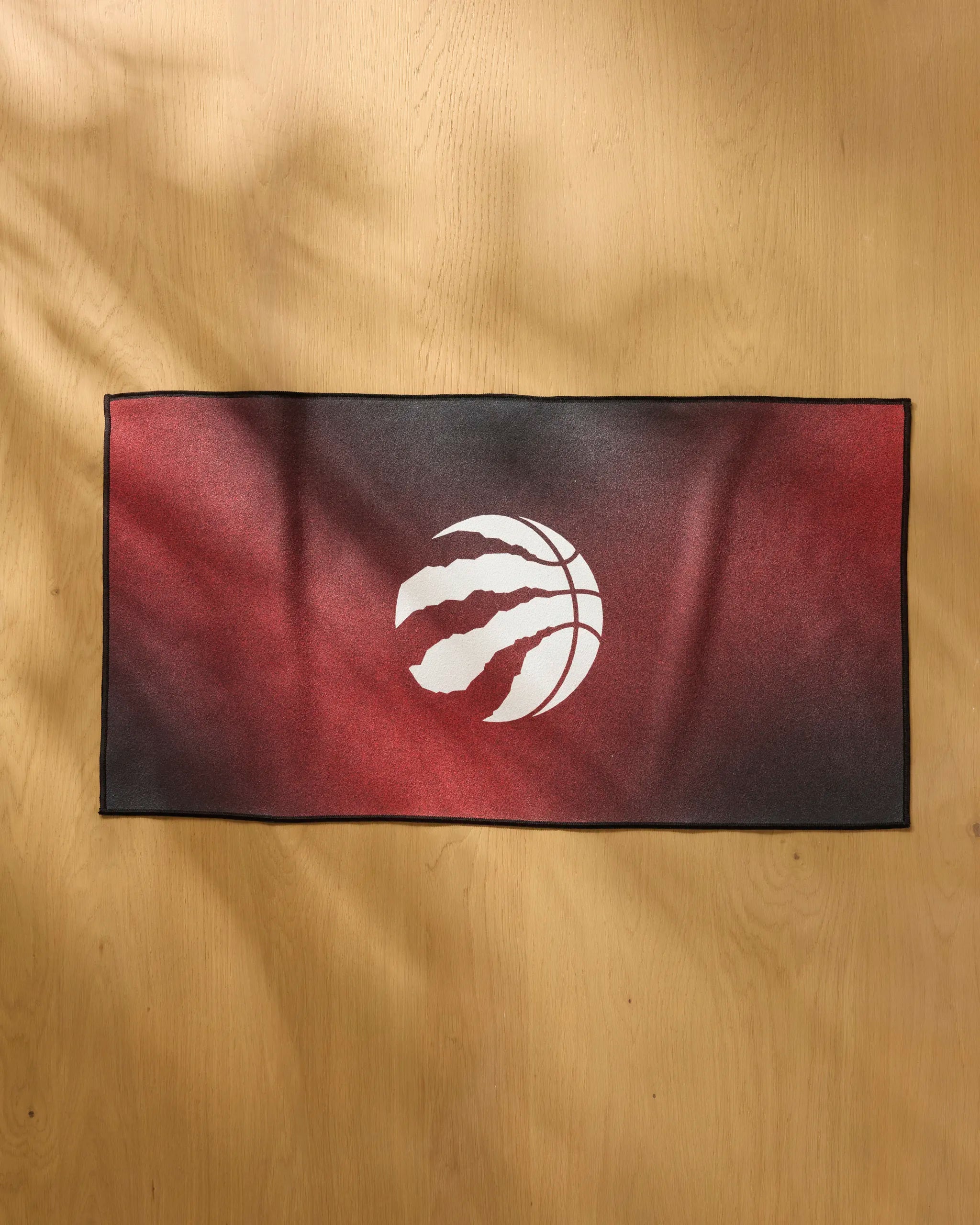 The NBALAB x Geometry Raptors Fade is a rectangular, quick-drying workout towel with a red and black gradient and a white basketball claw logo in the center, displayed on light wood.