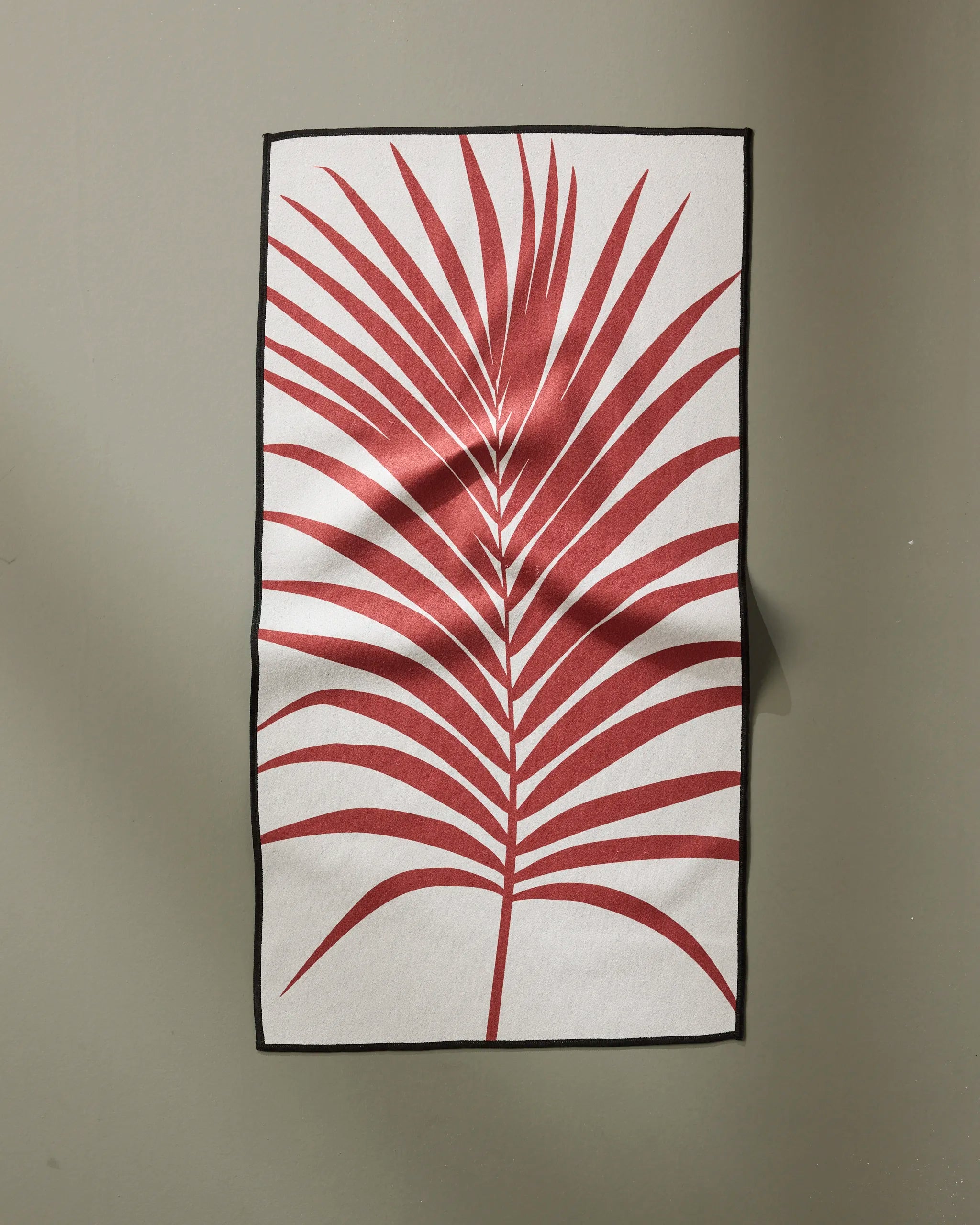 The PrintedPlant “Red Palm” is a rectangular white fitness towel made from recycled materials with a bold red palm leaf design at the center, a thin black border, and visible creases, shown hanging against a plain light gray wall.