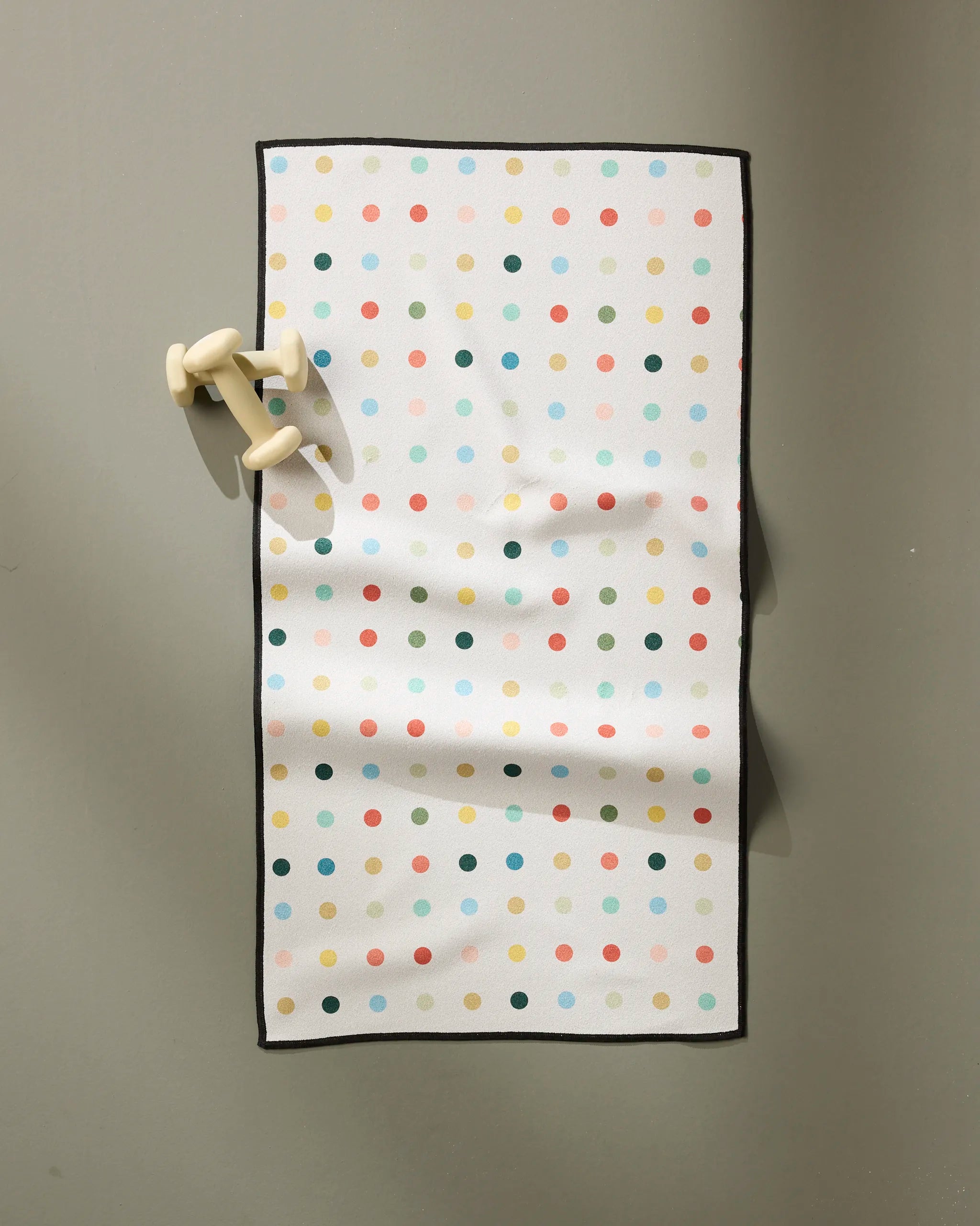 The Retro Dot fitness towel by Elizabeth Edwards is white, ultra-absorbent, rectangular, and made from recycled materials, featuring colorful grid polka dots, black edging, and hangs by beige hooks on a gray wall.