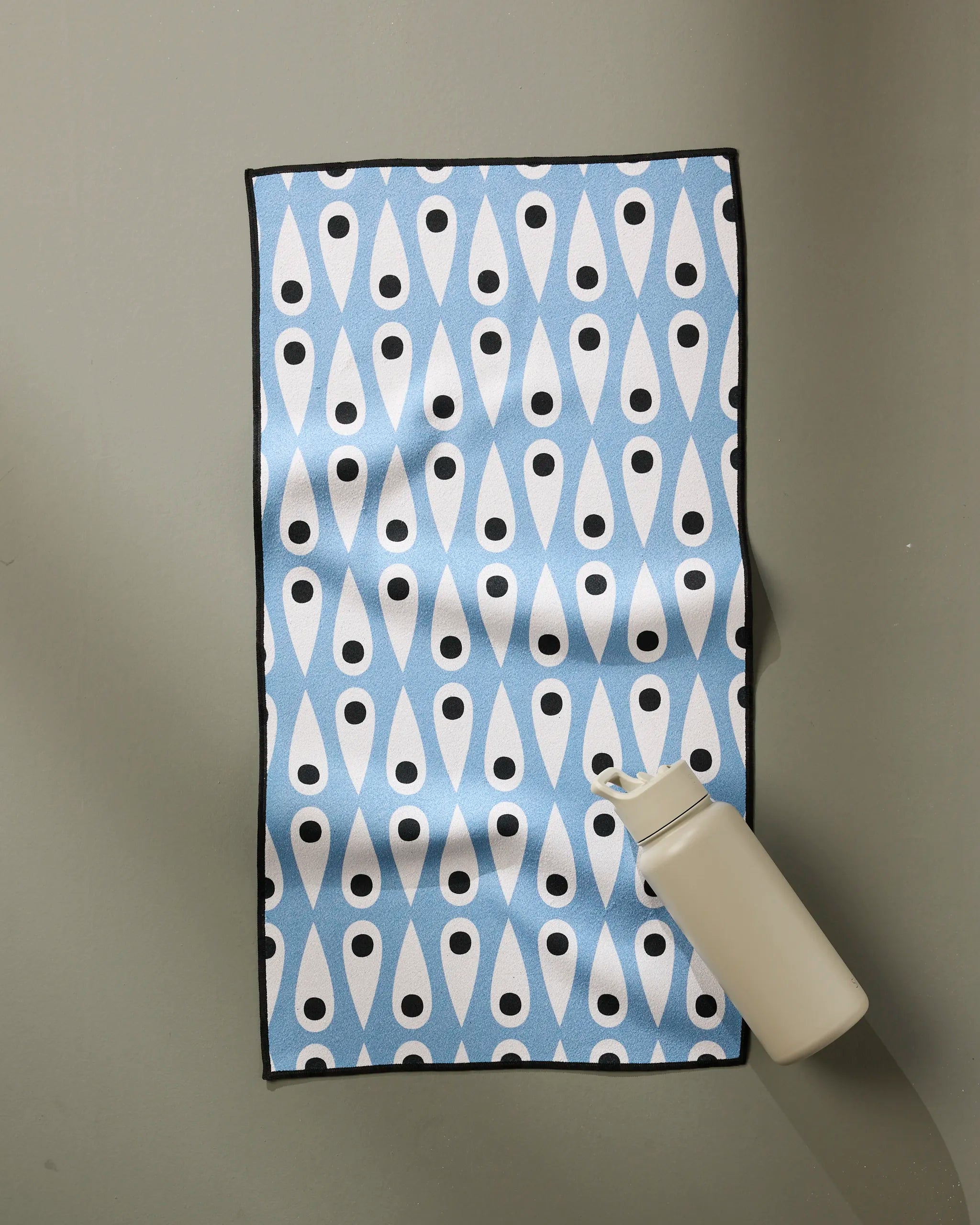 The Anette Heiberg Retro Rain fitness towel, in a light blue and white pattern with black dots, lies flat on a gray surface with a beige water bottle on one corner. Made from ultra-absorbent recycled materials for top performance.