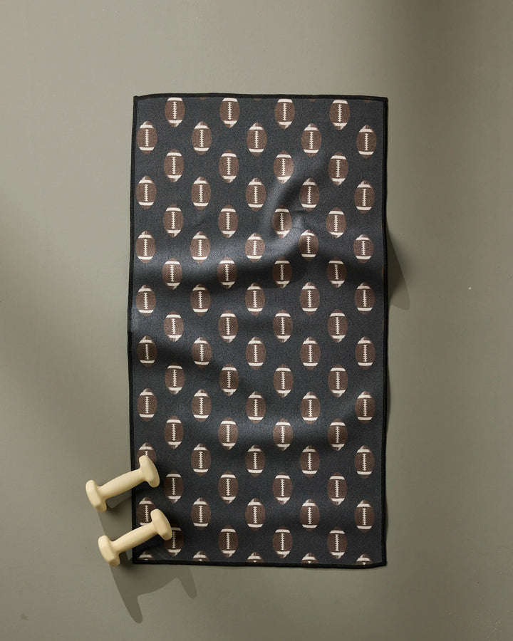 The Geometry Rush black fitness mat, made from recycled materials and featuring a repeating white “I” logo pattern, rests on a grey floor with two cream hand weights at the mat's bottom left corner.