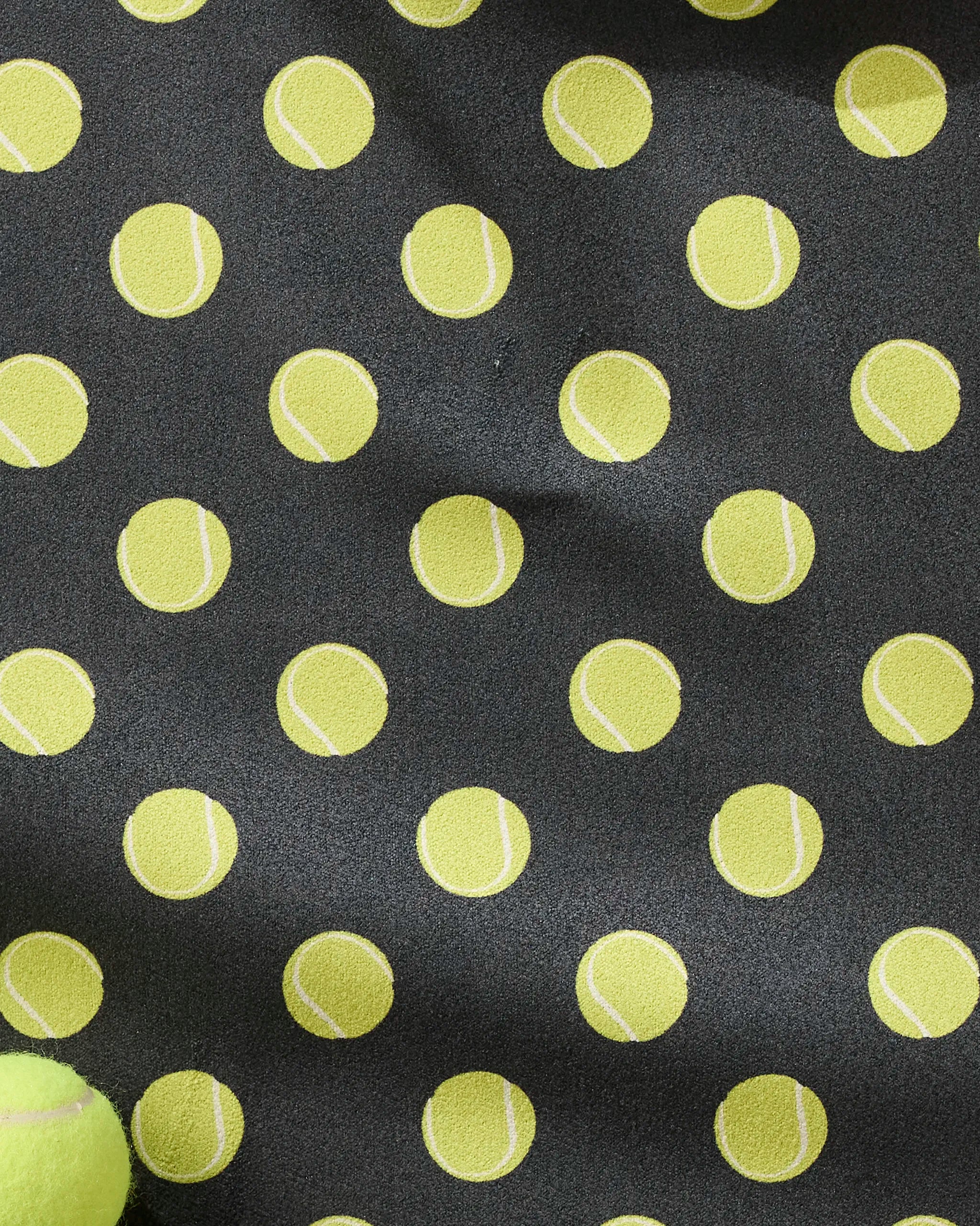 The Geometry "Serve" black fitness towel features evenly spaced yellow tennis balls with white curves, made from ultra-absorbent recycled materials. A real tennis ball appears in the lower left corner.
