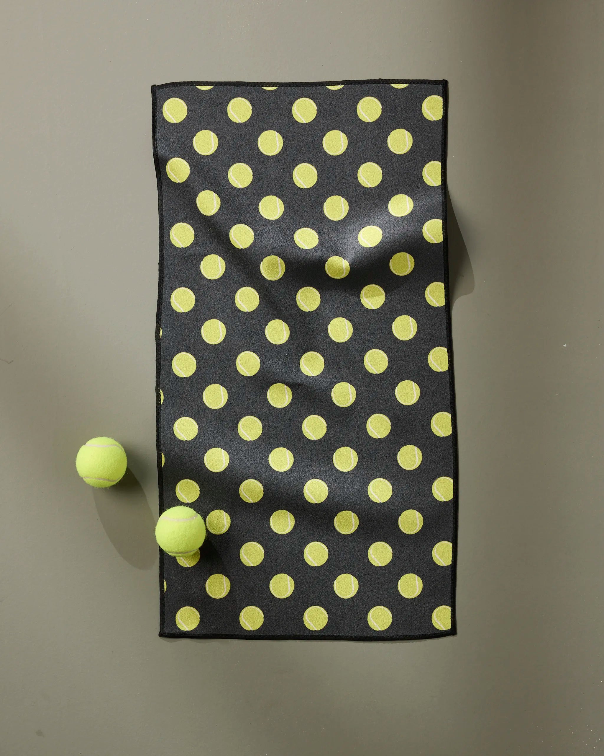 The Geometry Serve mat, a black rectangular mat made from recycled materials and patterned with yellow dots, rests on a gray surface with two yellow tennis balls placed beside it.