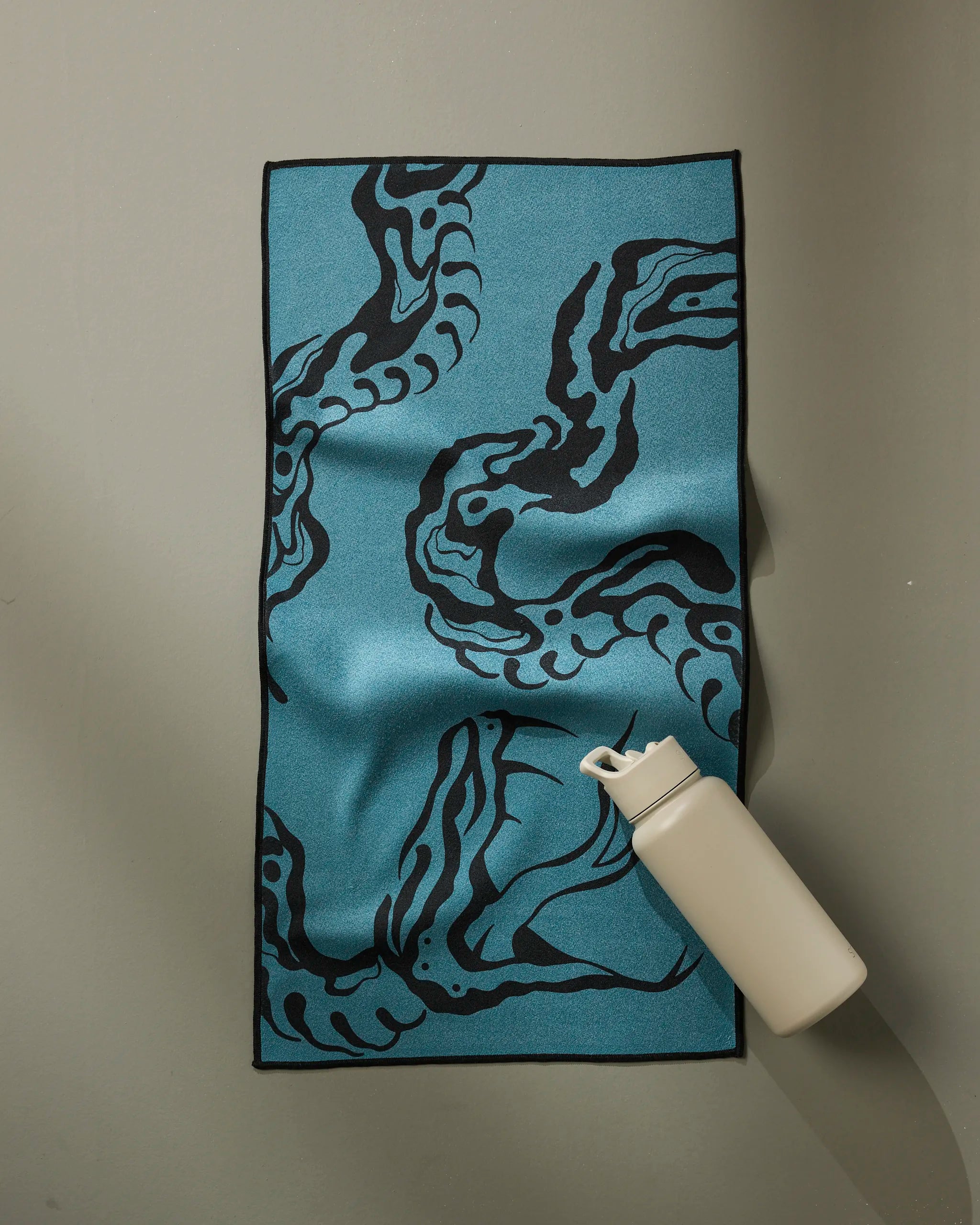 The Cole Strem "Snake" ultra-absorbent blue fitness towel with a black abstract tentacle design is on a gray surface beside an off-white reusable water bottle.
