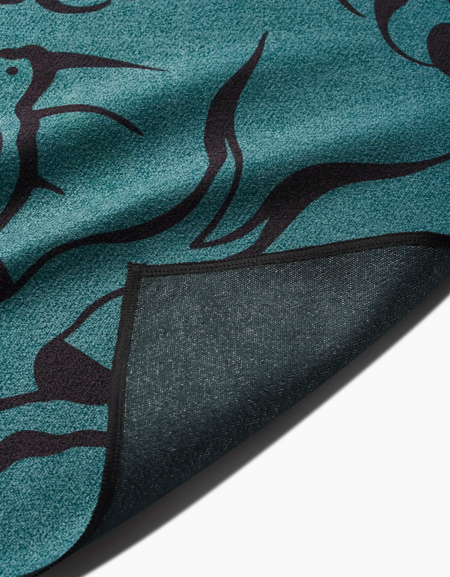 A close-up of the Cole Strem Snake rug in teal and black shows its abstract design with a flipped corner revealing the underside. Made from recycled materials, the rugs texture is clearly visible.