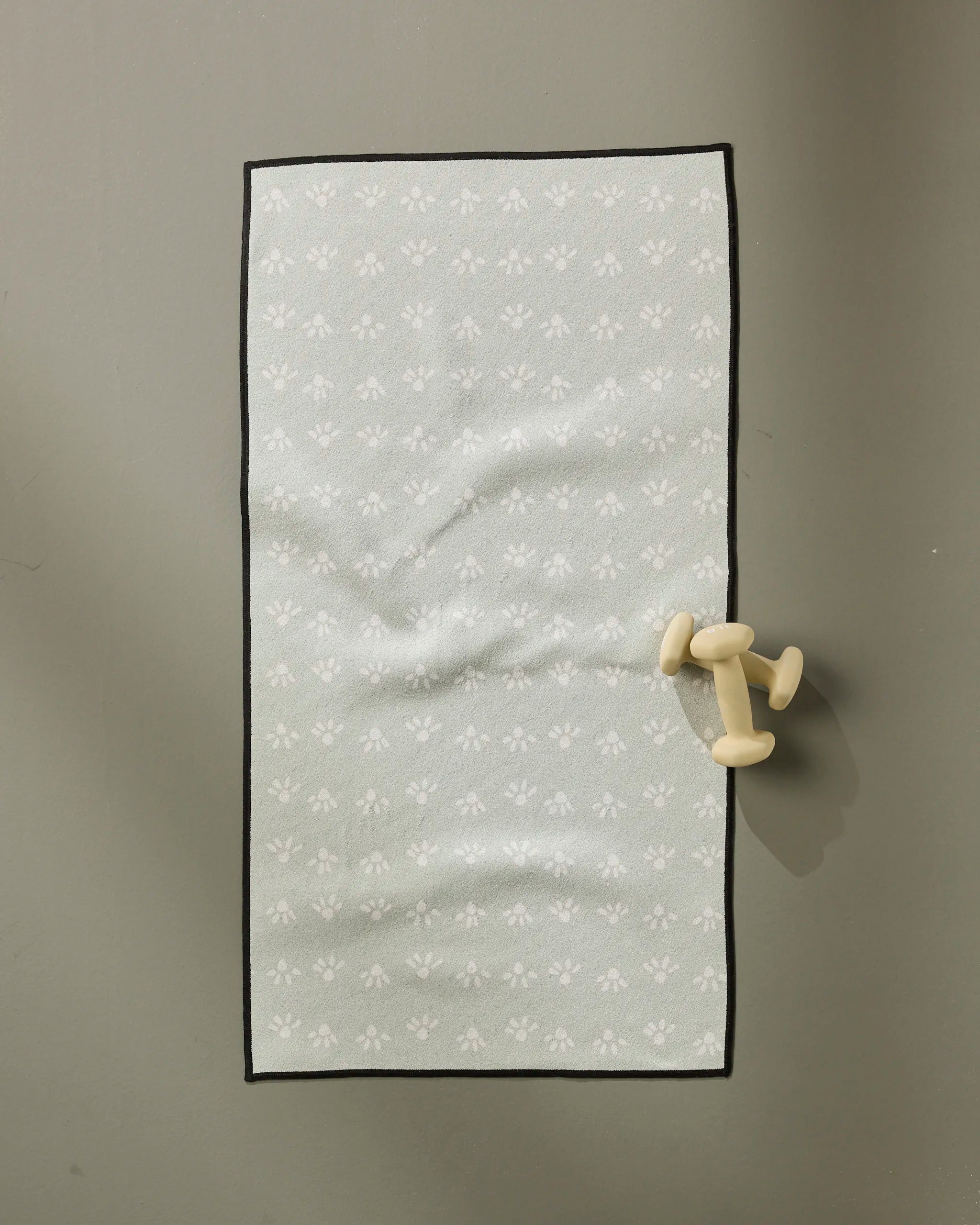 A single small dumbbell rests beside an ultra-absorbent towel on the "Sun Bursts" yoga mat by Anee Shah, featuring light gray with subtle white floral designs, placed on a smooth gray surface.