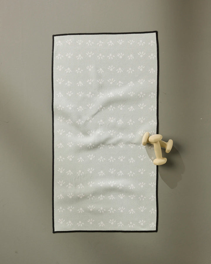 A single small dumbbell rests beside an ultra-absorbent towel on the "Sun Bursts" yoga mat by Anee Shah, featuring light gray with subtle white floral designs, placed on a smooth gray surface.