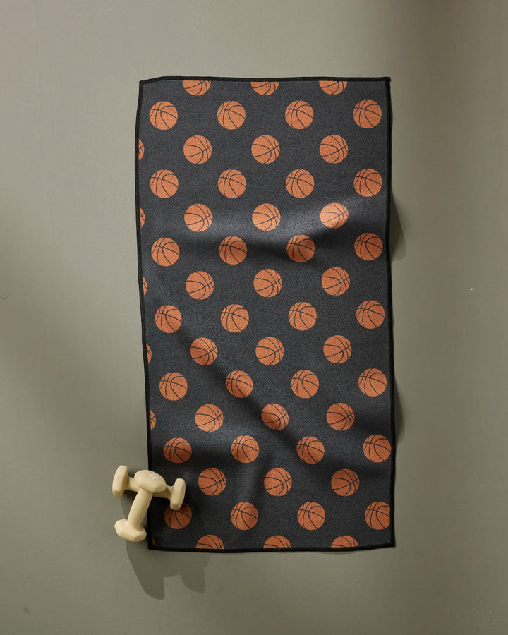 The Geometry Swish fitness towel, featuring an orange basketball pattern on black and fast-drying fabric, hangs on a gray wall with a small dumbbell resting nearby.