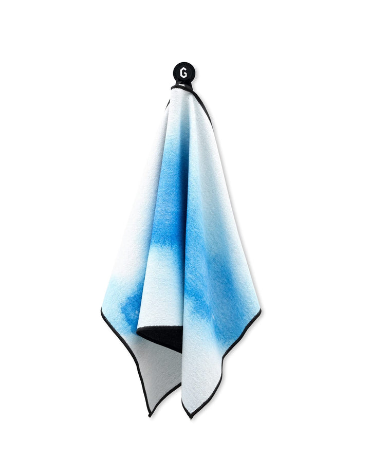 A Magna - The Soul towel by GEOMETRY with a blue gradient hangs from a black hook on a white background. Featuring black edging, this gym towel is ultra absorbent, soft, and ideal for any fitness routine.