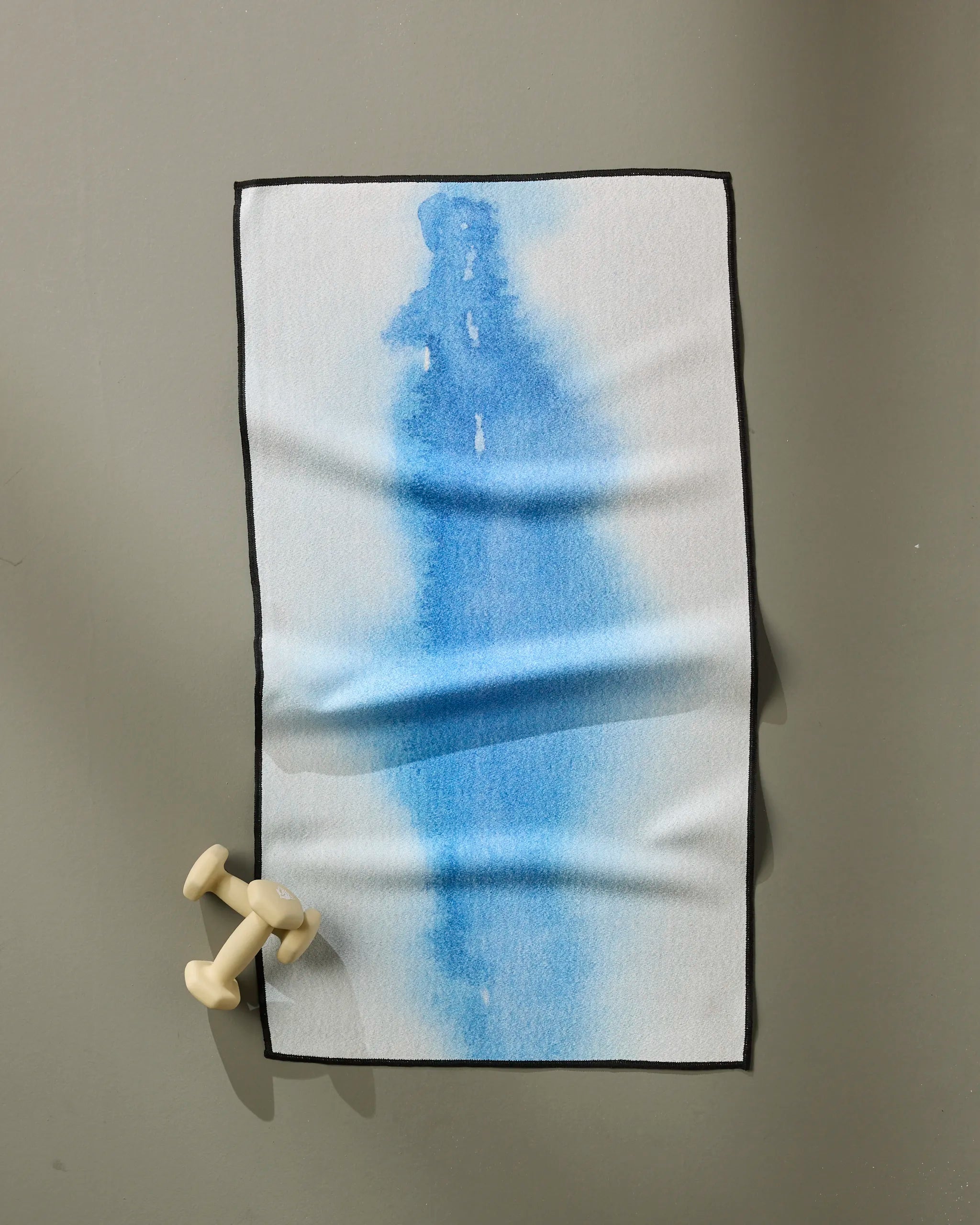 The Soul fitness towel by GEOMETRY, ultra-absorbent and small with a blue water stain design, hangs on a neutral wall above a light beige dumbbell.
