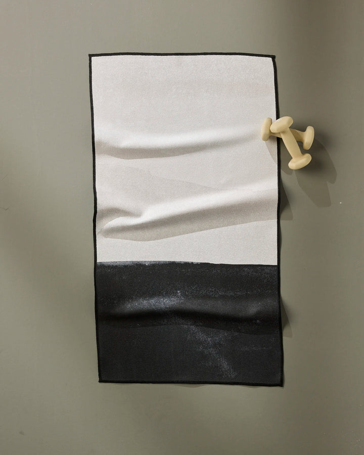 The Top Layer fitness towel by GEOMETRY features a sleek black and white color block design, displayed on a dumbbell-shaped modern hook. Crafted from ultra-absorbent, recycled materials for eco-friendly performance.