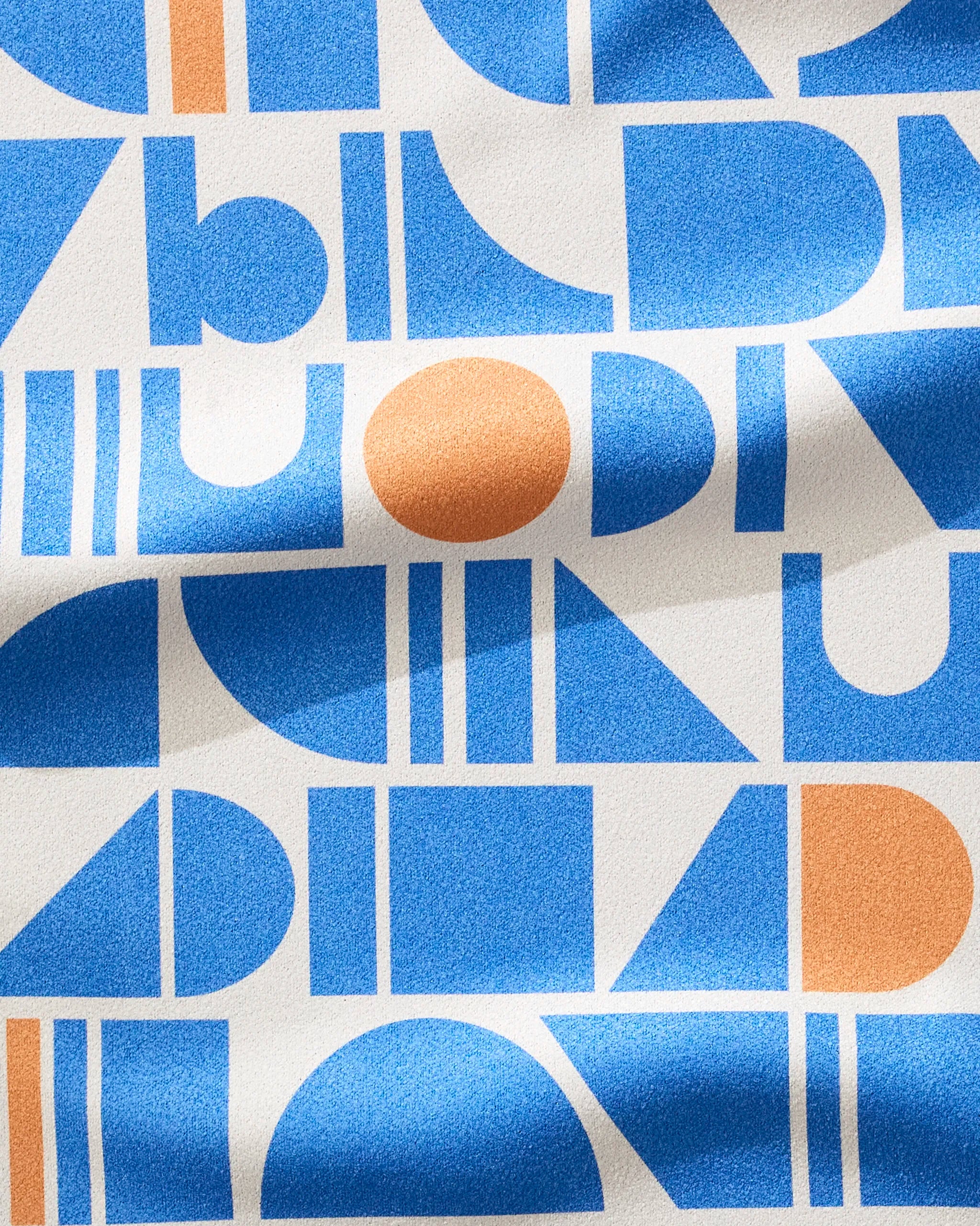 Ultra Marine Harmony by Happy Hemera is an eco-friendly fabric made from recycled materials, featuring blue and orange geometric shapes—circles, triangles, rectangles, semicircles—on a white grid with folds and shadows for depth.