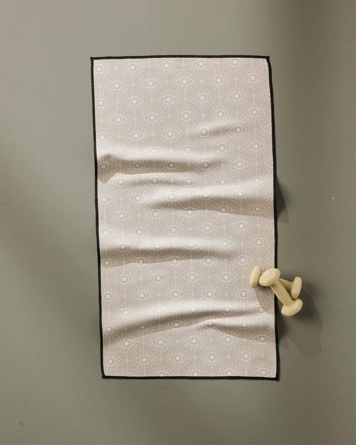 The Geometry Vision towel is a rectangular, light pink fitness towel with a subtle eye pattern. Ultra-absorbent and quick-drying, it hangs on a gray wall from a cream-colored, abstract hook shaped like bent legs.