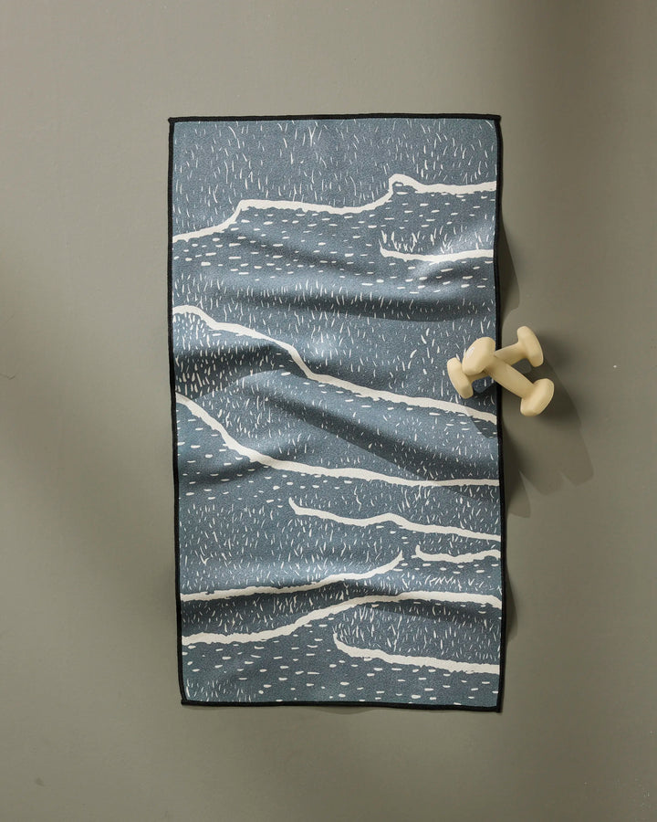 The GEOMETRY Wetlands Midnight fitness towel, featuring blue with white wavy lines, is spread on a gray floor with light-colored dumbbells placed on its right edge.