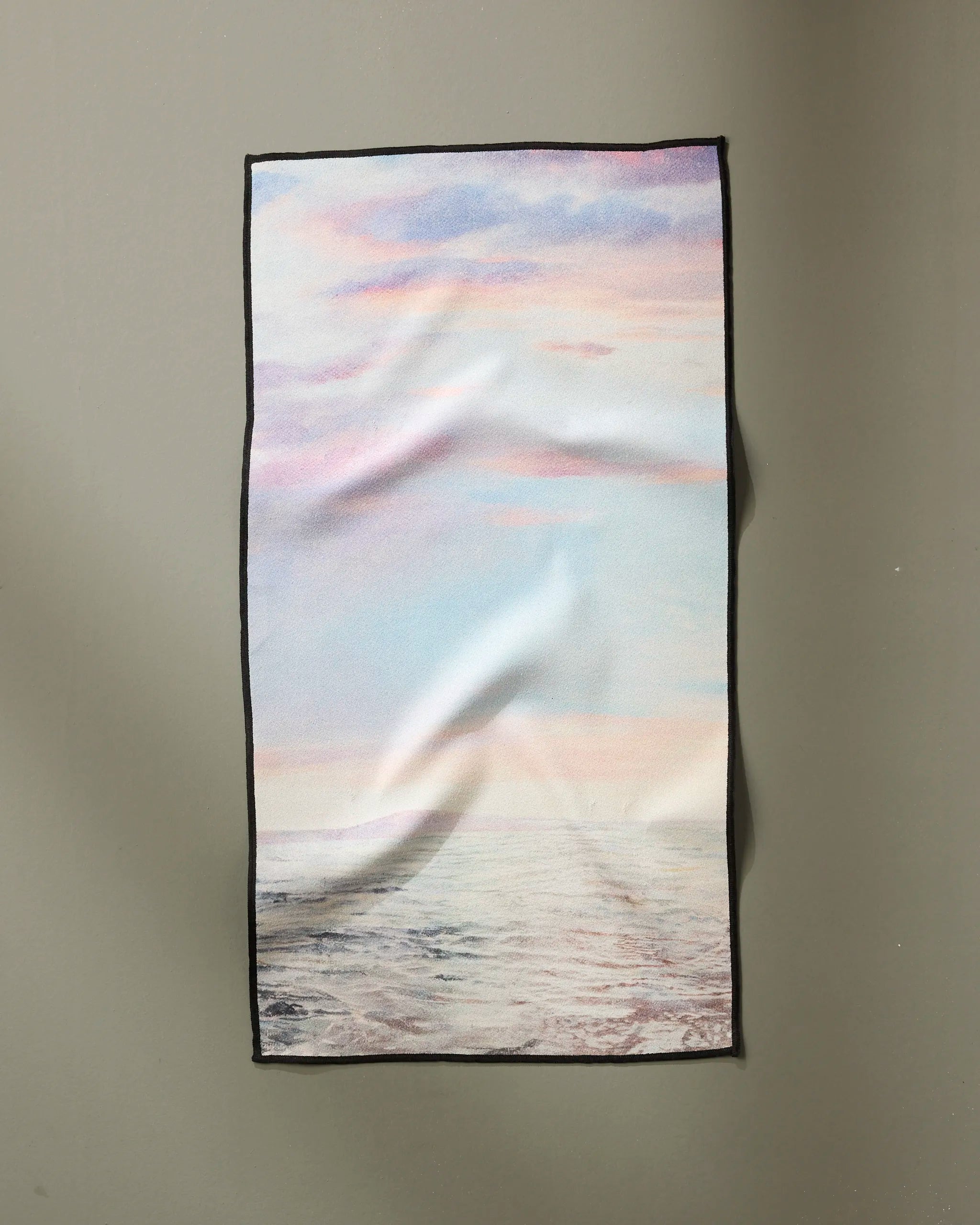 “When I Close My Eyes” by Stefanie Bales is a rectangular, ultra-absorbent cloth featuring a printed calm sea and pastel sky with clouds, bordered in black and displayed against a plain gray wall.