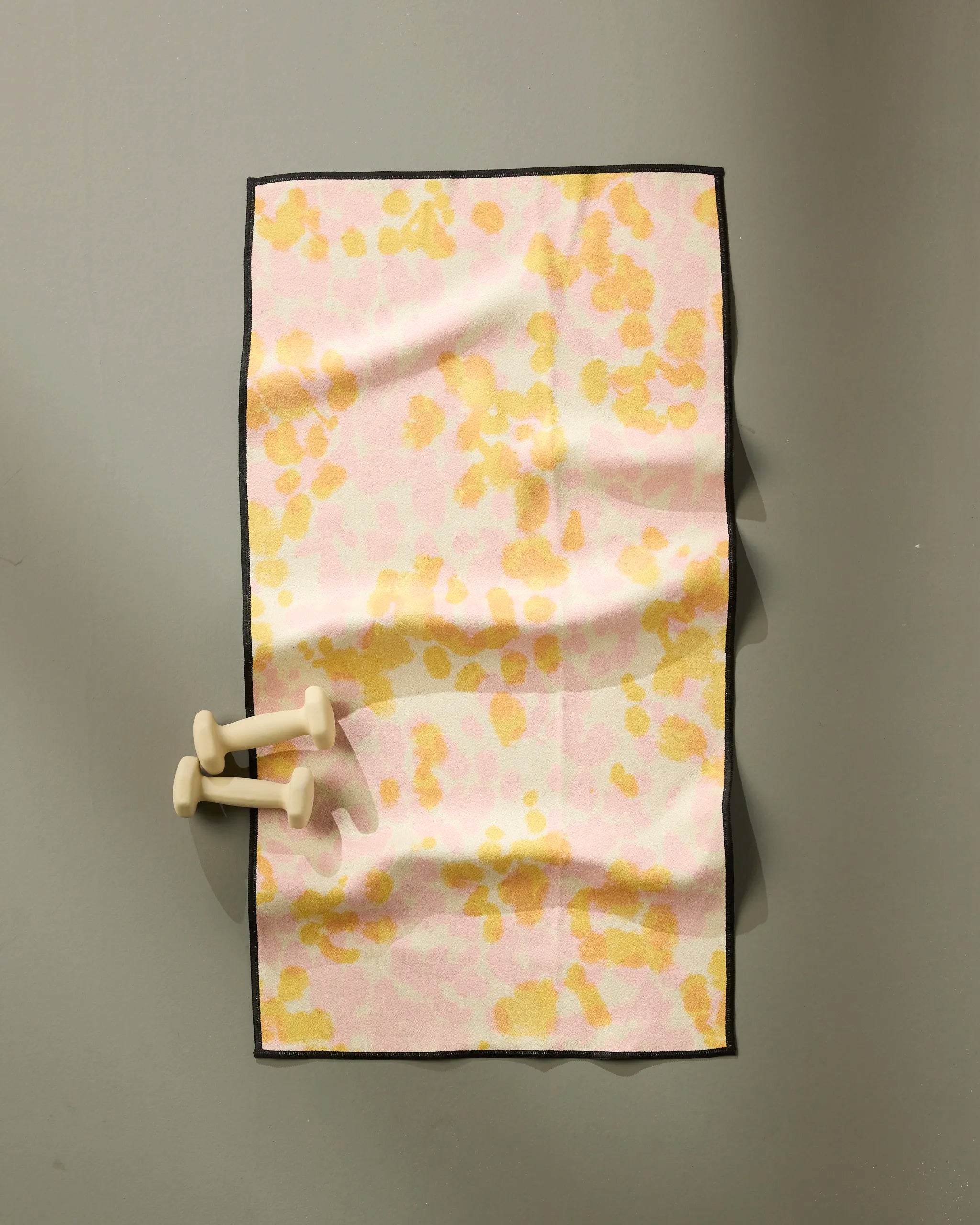 The Whimsy exercise mat by GEOMETRY features a pink and yellow abstract pattern with black edging, is made from recycled materials, and is shown on a gray surface next to two light beige dumbbells.