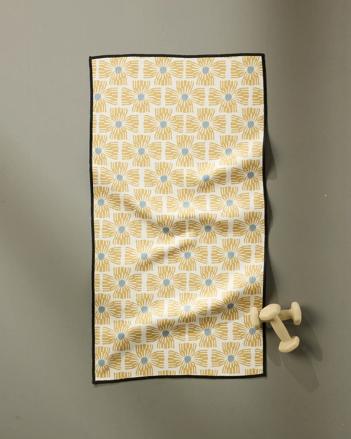 The Ash Cascade Wildflower fitness towel, ultra-absorbent and made from recycled materials, features a yellow and blue geometric pattern with a black border. It hangs on a gray wall with light-colored dumbbells resting on one end.