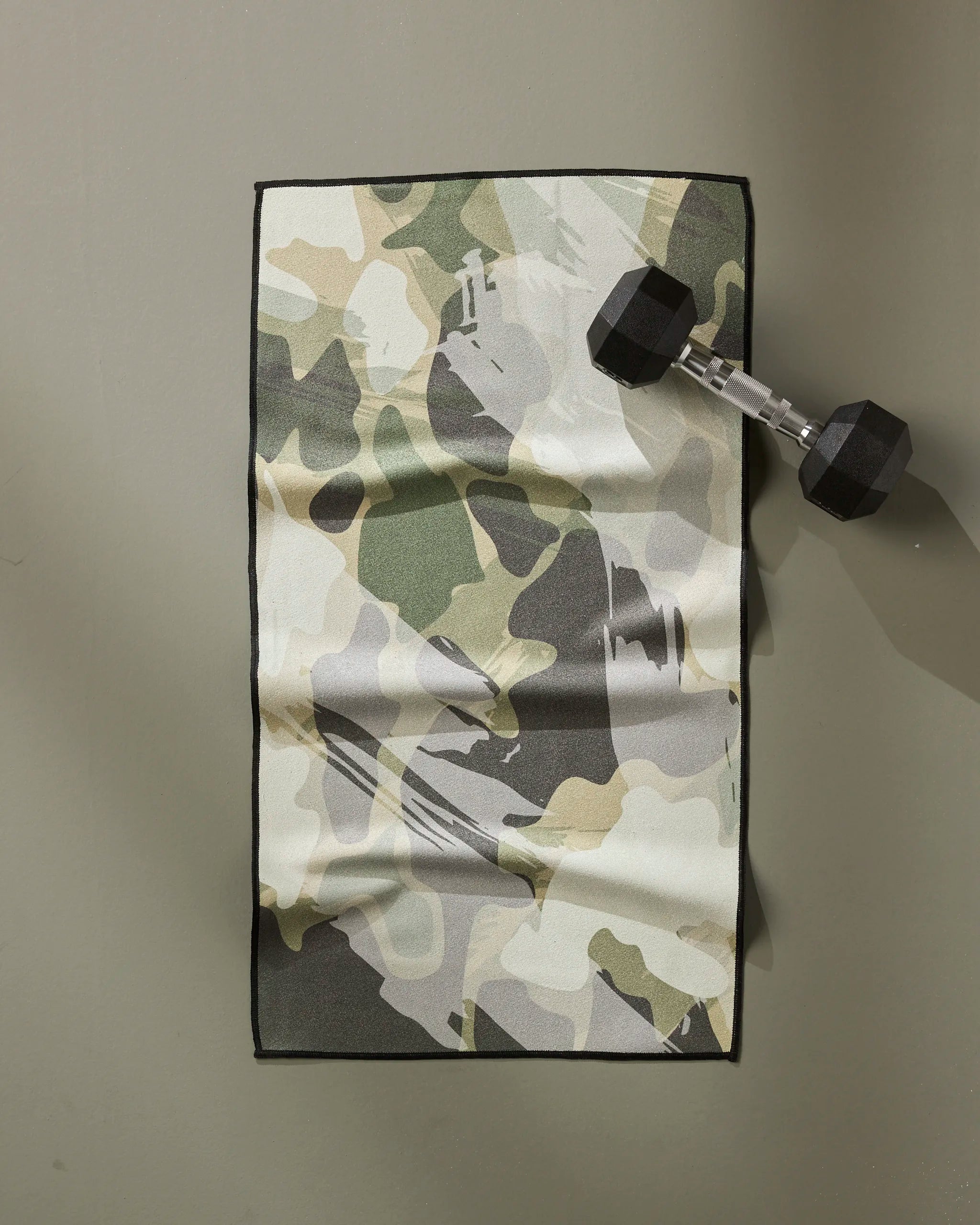 The GEOMETRY Wildscape towel, featuring a camouflage pattern and made from recycled materials, is shown on a light green surface with a black dumbbell on its top right corner.