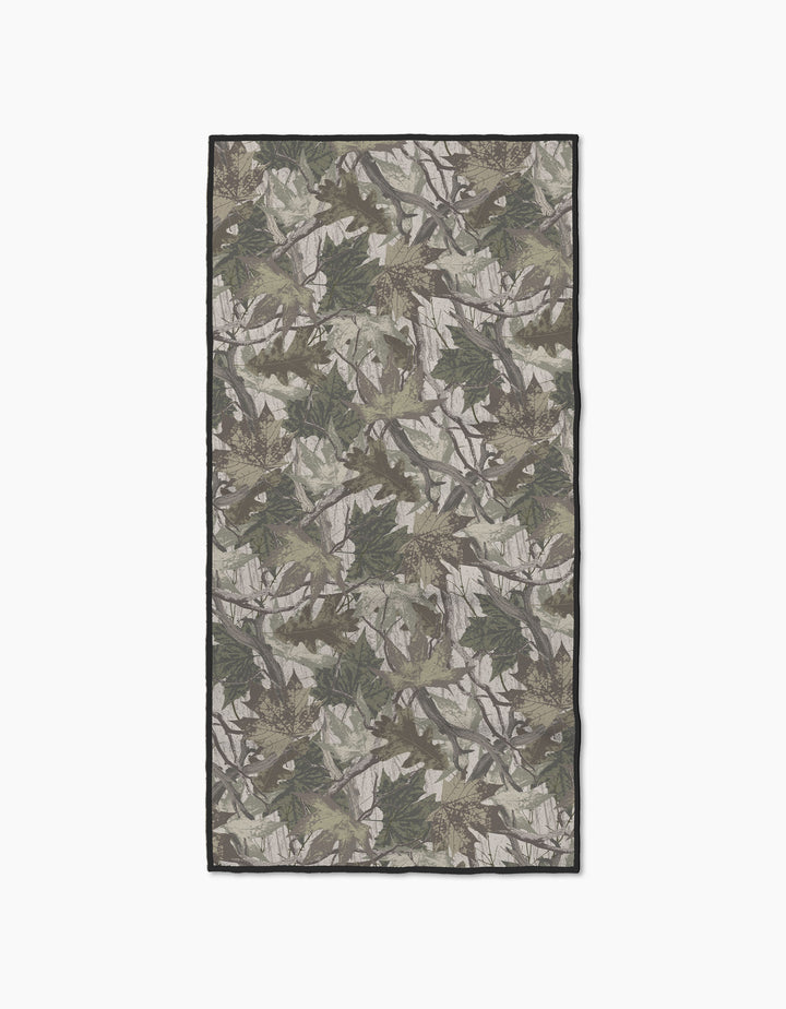 The Geometry Dad Camo fitness towel is rectangular, made from recycled materials, and showcases a green, brown, and gray camouflage pattern with leaf and branch designs on a plain white background.