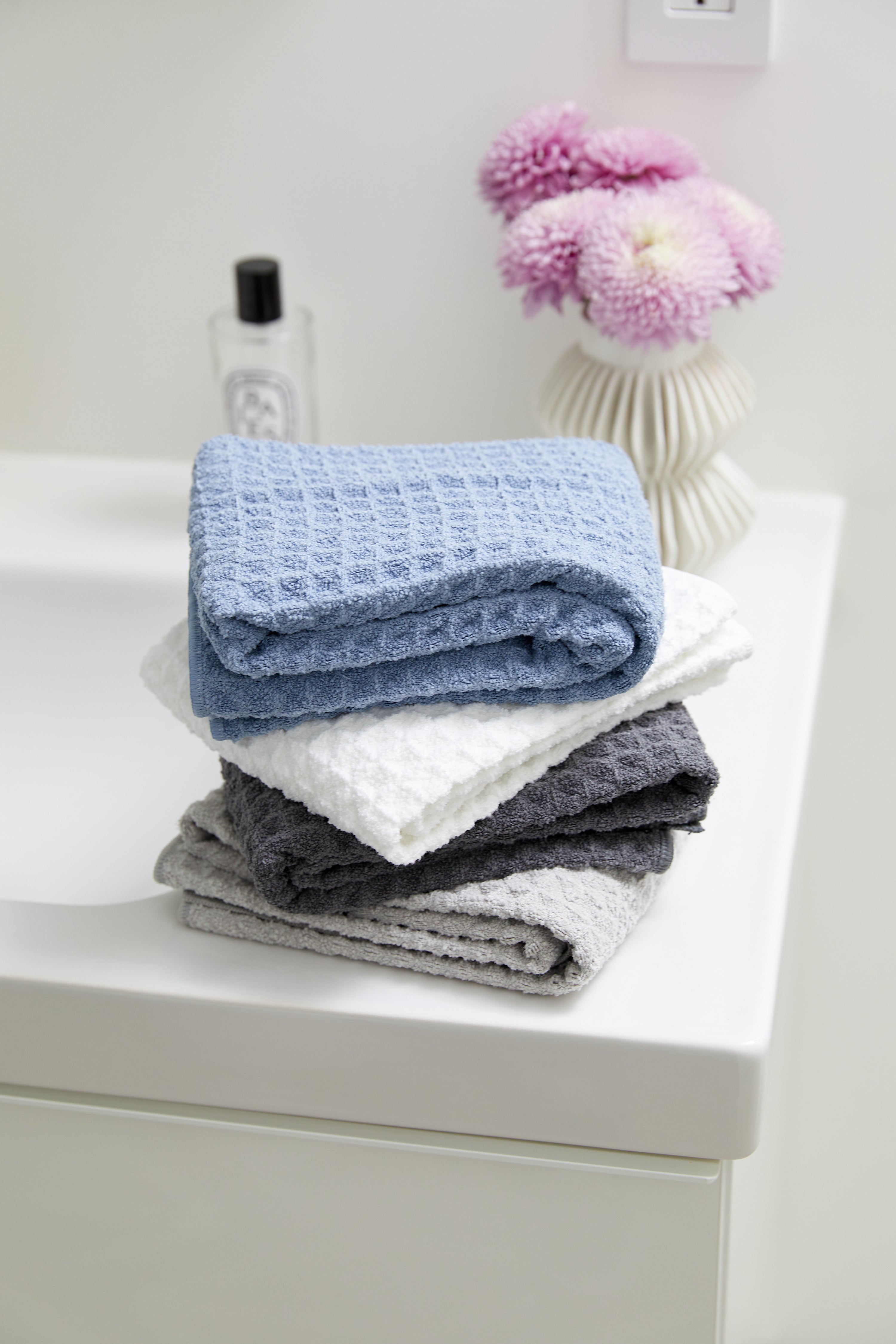 A stack of four GEOMETRY Cloud luxury waffle hand towels in blue, white, dark gray, and light gray rests on a white bathroom counter with a white vase of pink flowers and a clear bottle in the background.