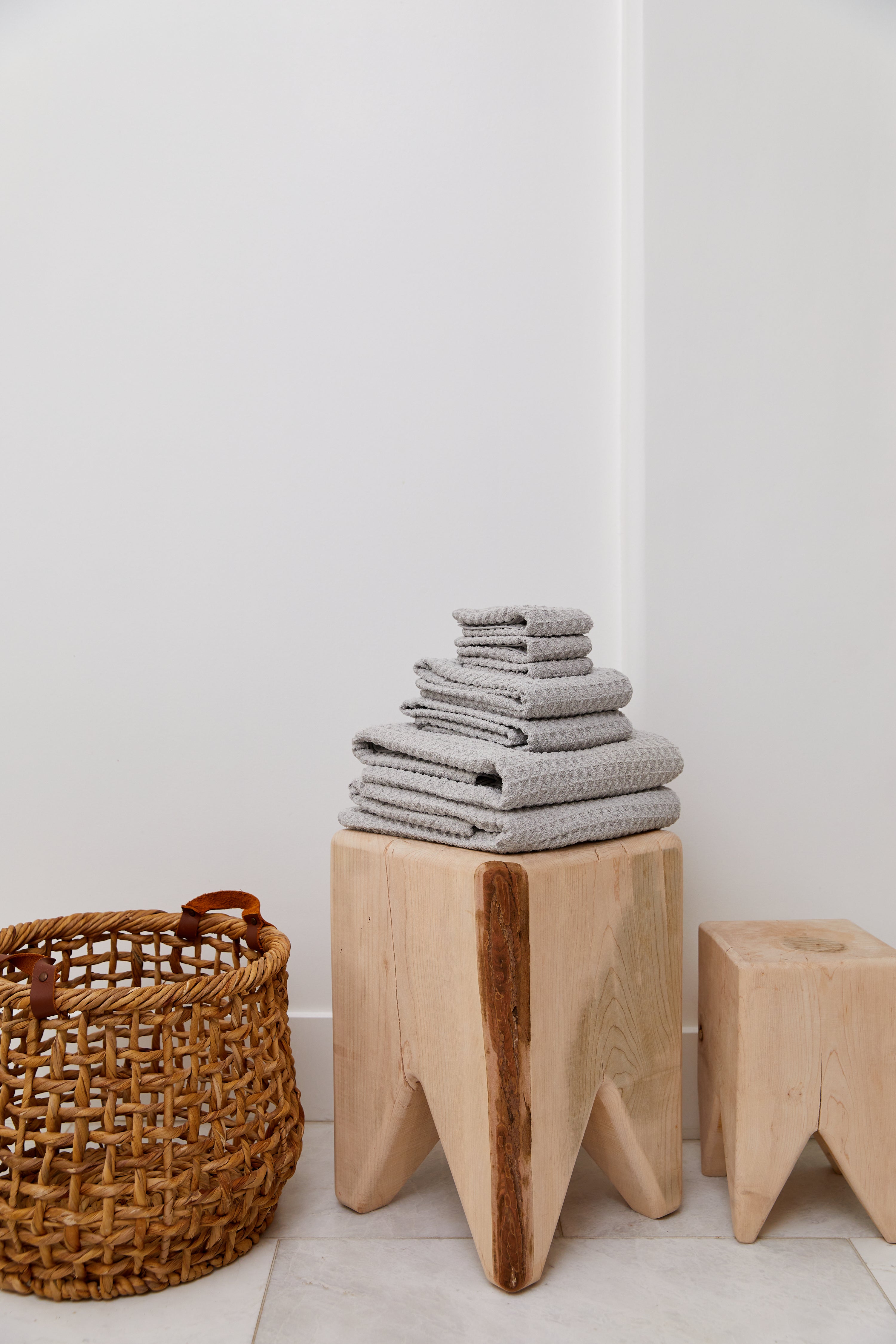 A stack of GEOMETRY Stone luxury towels sits on a light wooden stool beside a woven basket and a smaller wooden stool, set against a white wall and tiled floor.