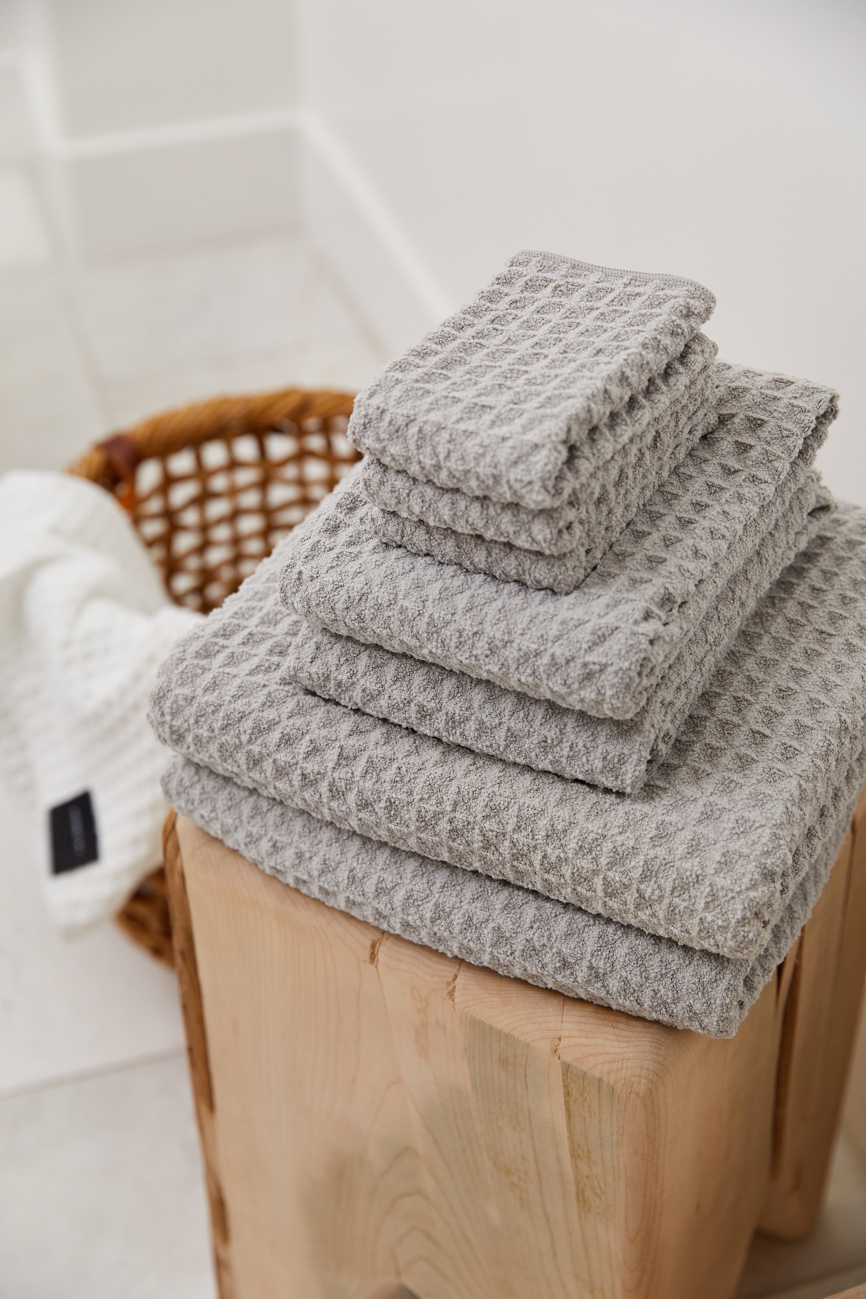 A neatly stacked set of GEOMETRY Stone gray ultra-absorbent Waffle Bath Towels sits on a wooden stool, with a wicker basket holding a white towel in the background on a tiled floor.