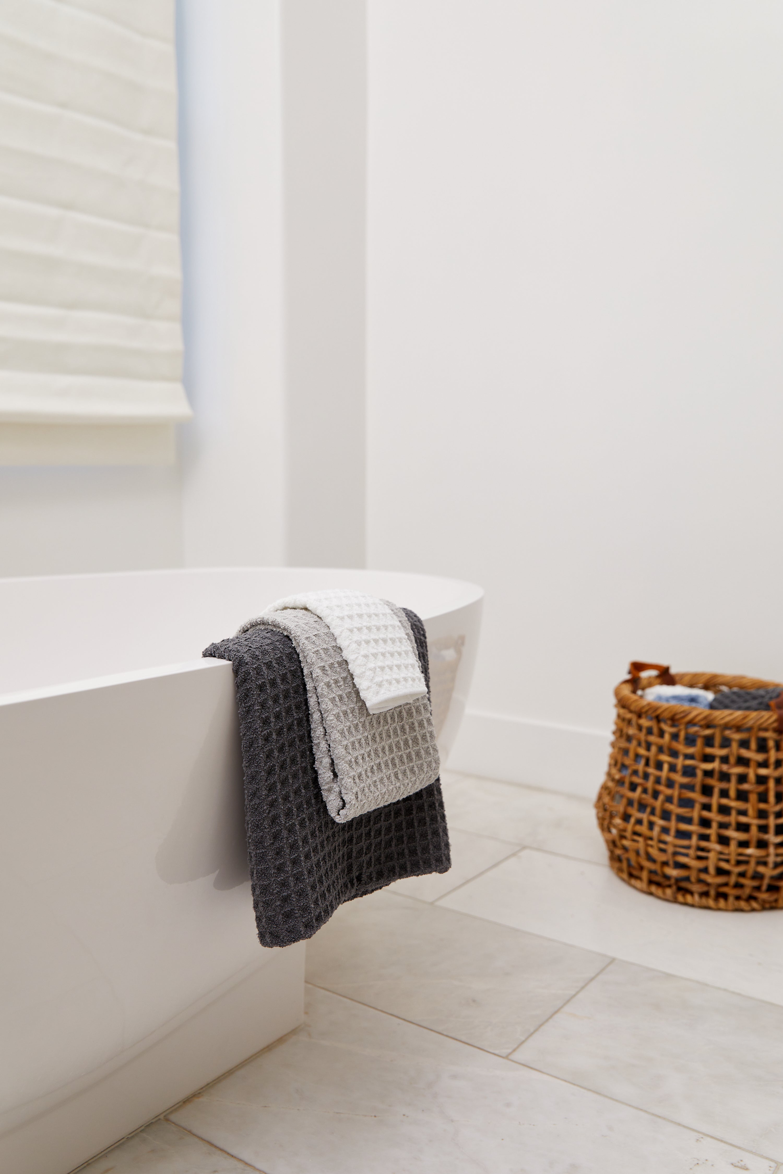 A modern bathroom features the GEOMETRY Cloud white bathtub, adorned with luxurious textured gray and white towels. A wicker basket of towels sits on light tiles near a white wall and shaded window.