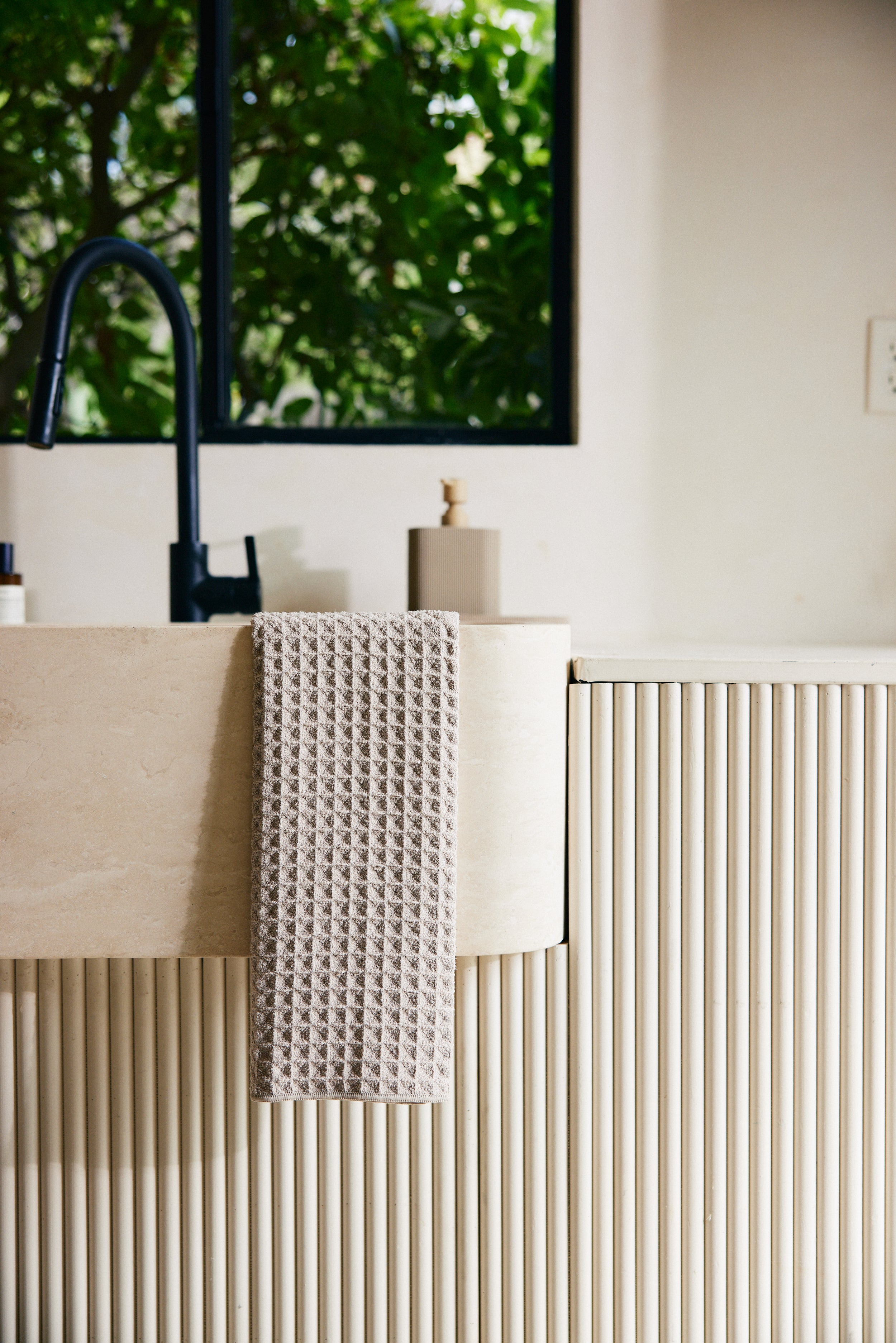 A GEOMETRY Taupe waffle hand towel hangs over a cream-colored, vertically-grooved sink next to a black faucet, with a soap dispenser and a window showing green foliage in the background.