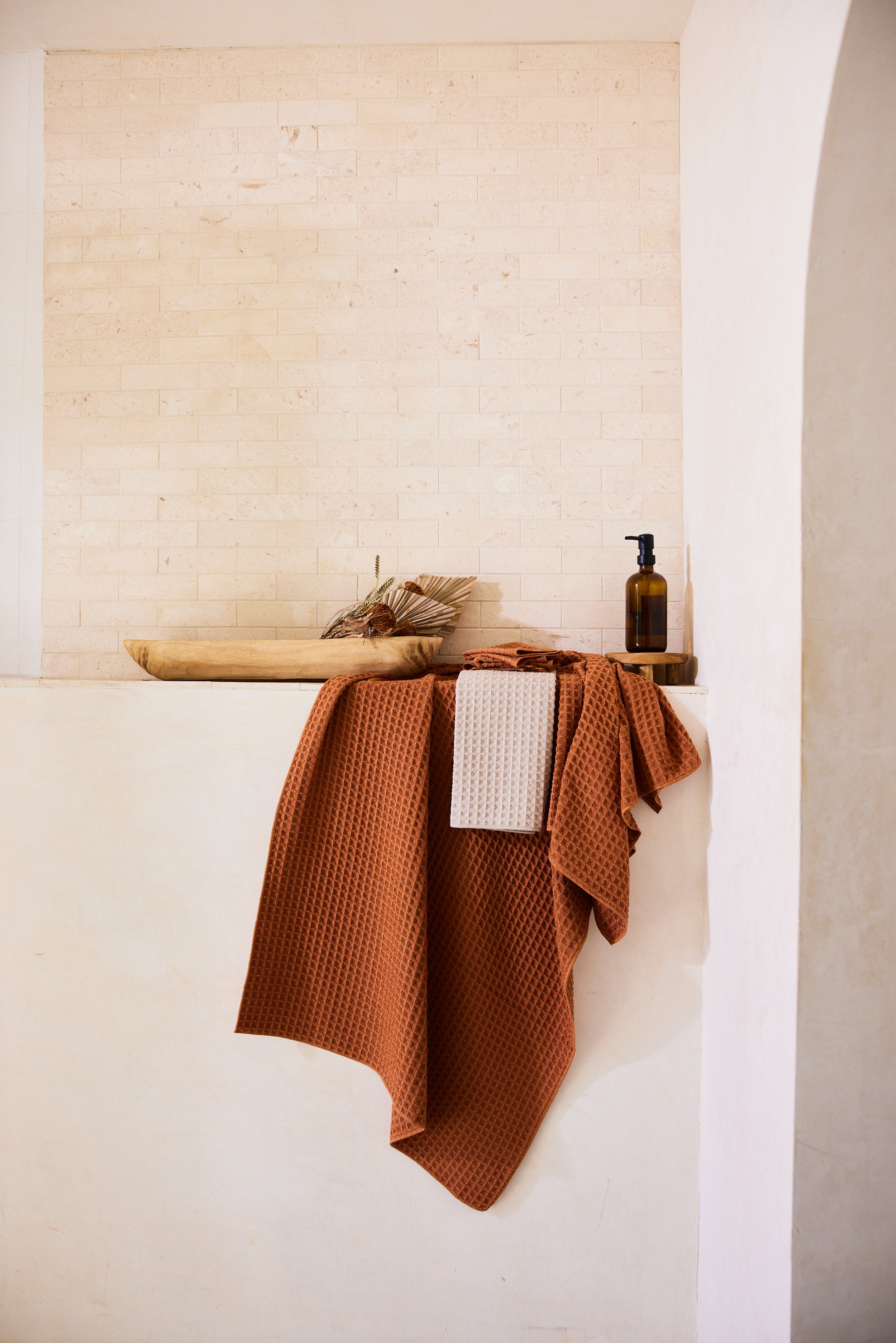 A Clay towel from GEOMETRYs Waffle Bath Collection and a beige cloth hang over a ledge in a minimalist bathroom with cream walls. On the ledge are a wooden tray, dried leaves, and an amber pump bottle.