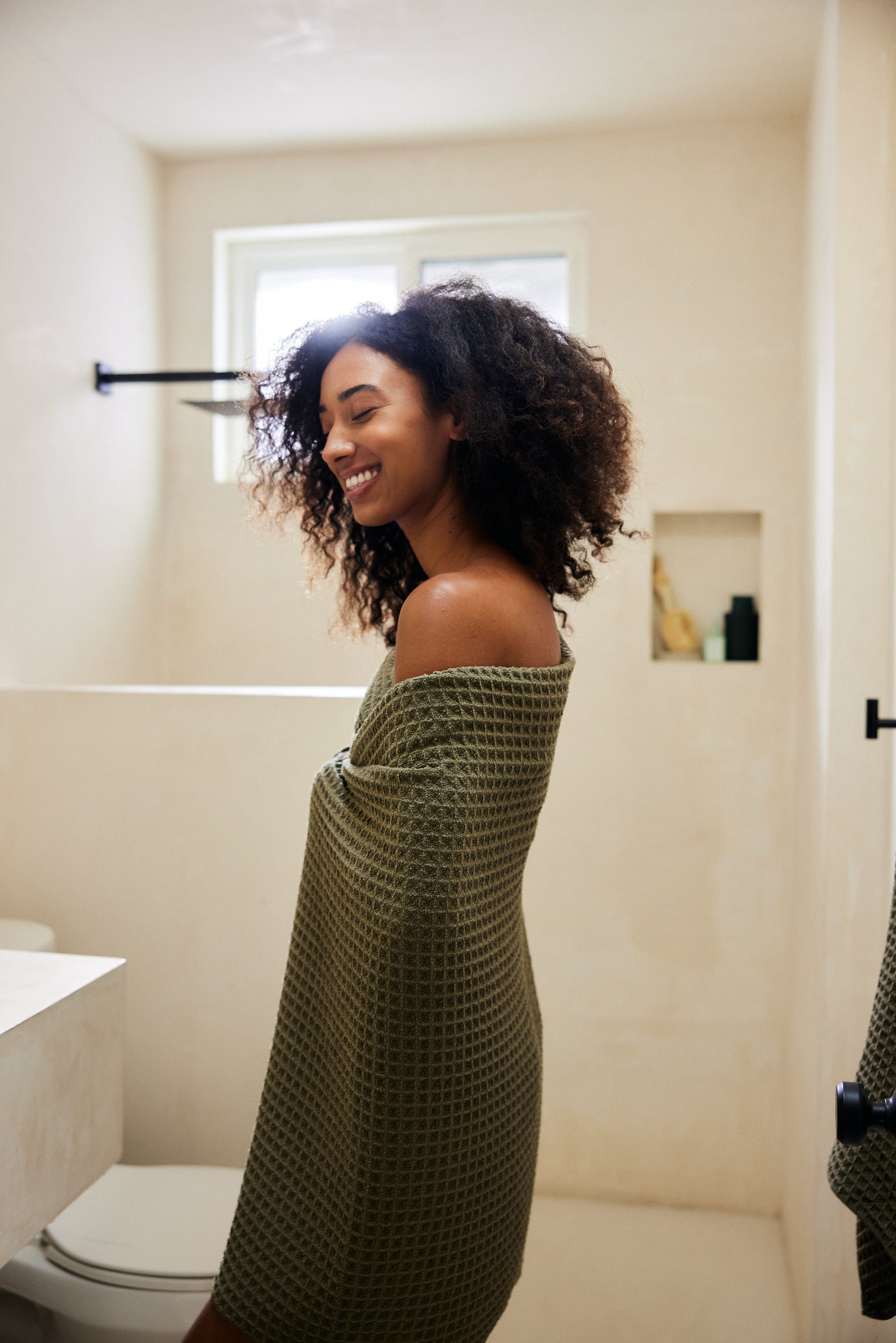 A woman with curly hair smiles in a sunlit bathroom, wrapped in the GEOMETRY Forest textured green waffle bath towel, with eco-friendly Forest towels stacked neatly on a shelf behind her.
