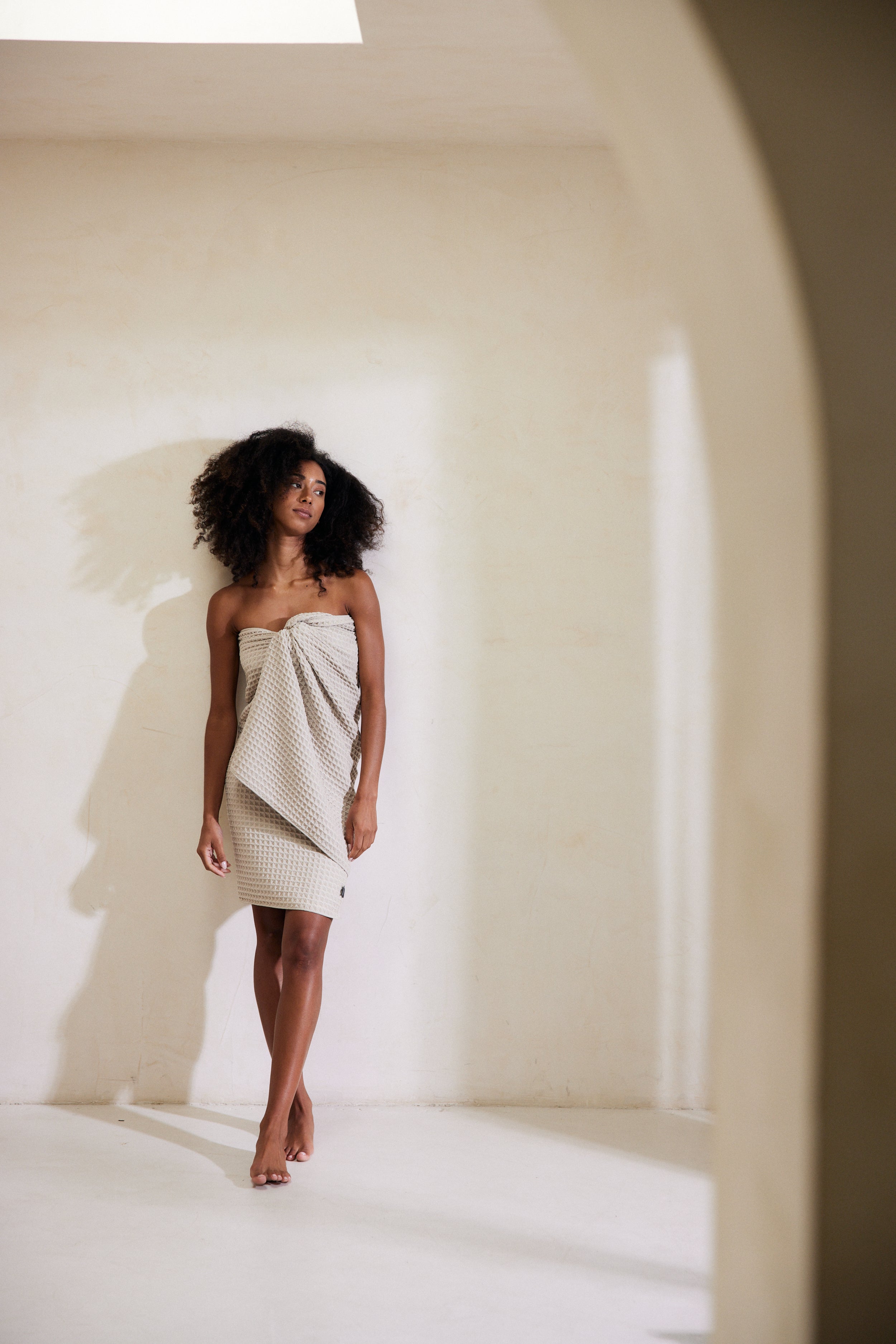 A woman with natural curls stands barefoot in a bright, minimalist room, wearing GEOMETRYs Taupe, a strapless textured cream dress made from recycled materials. She gazes to the side, casting a gentle shadow on the wall.
