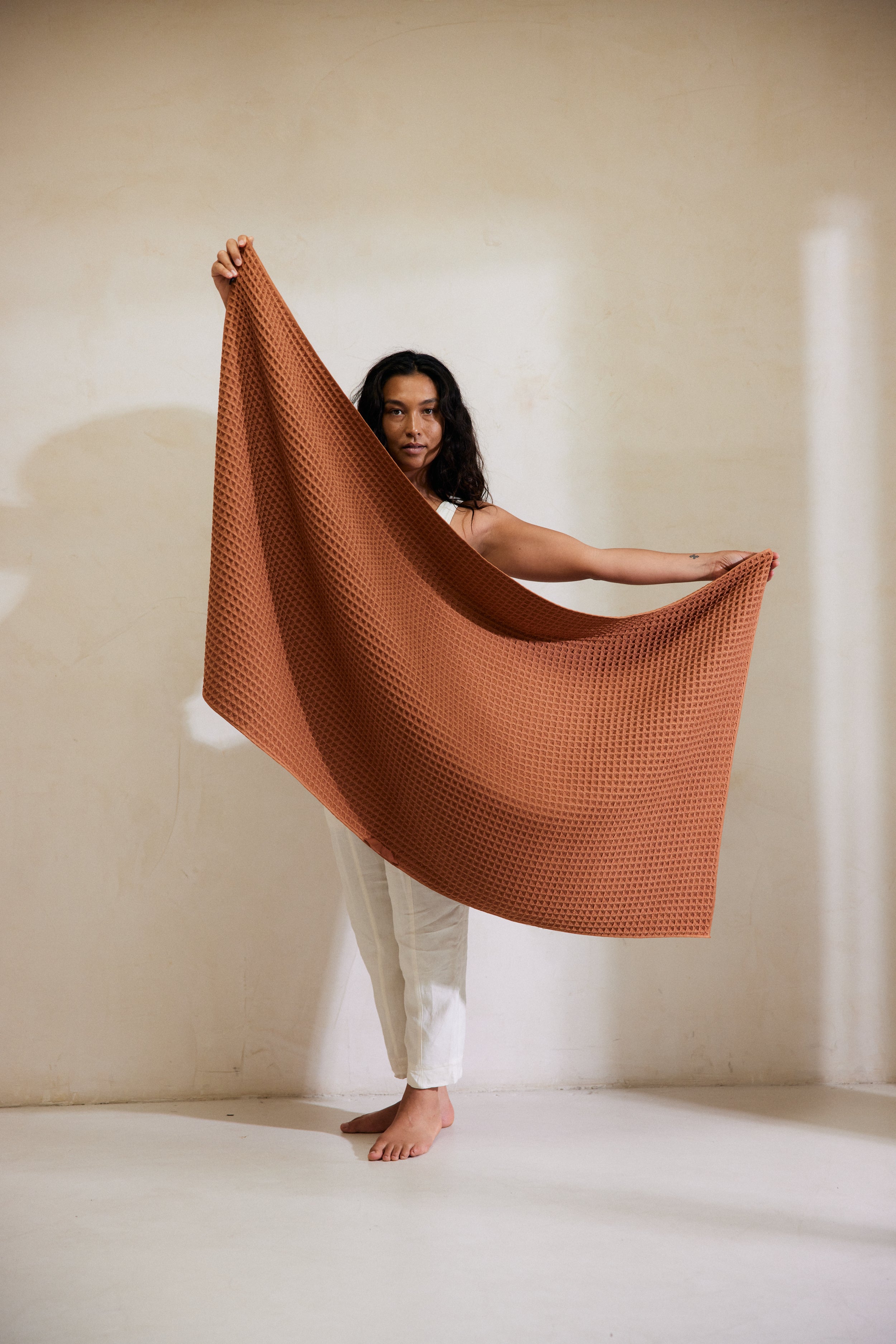 A person with long, dark hair and white pants stands barefoot on a white floor, holding GEOMETRYs Clay Waffle Bath Towel wide open in front of a light beige wall.