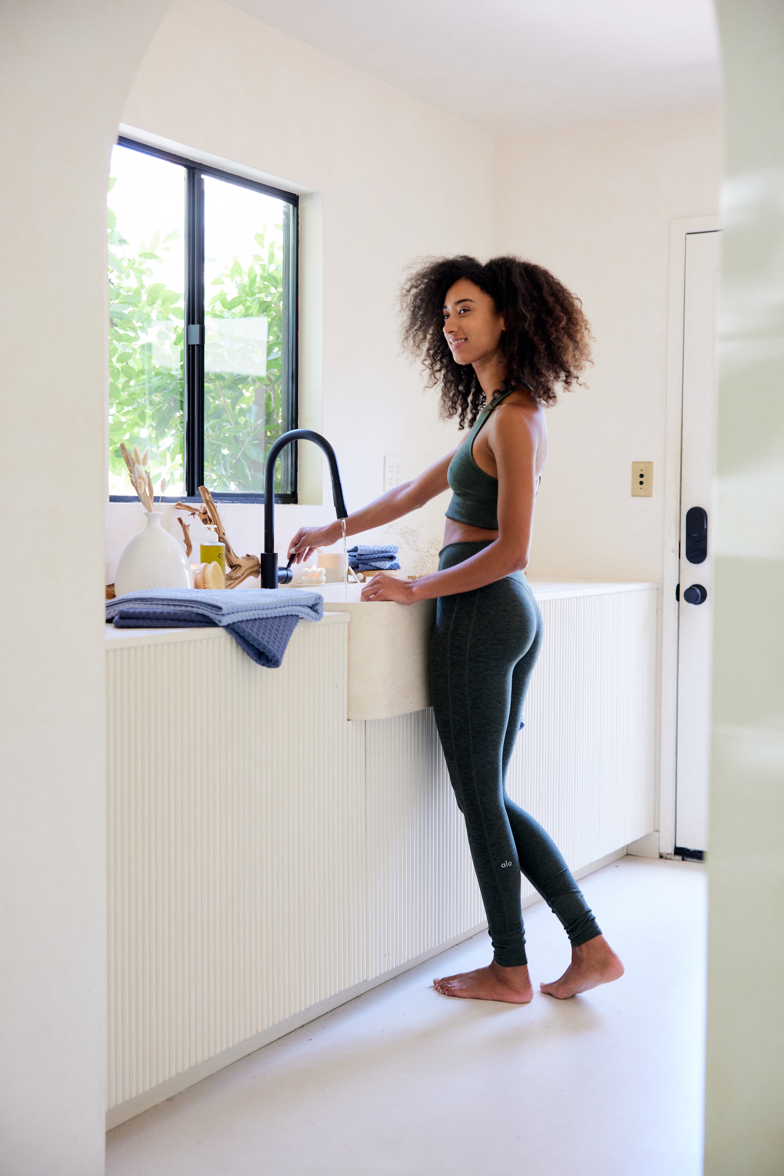 A woman in green activewear smiles as she washes dishes barefoot at a sunlit kitchen sink. On the counter are eco-friendly GEOMETRY towels in Midnight Blue, along with sustainable utensils. Green foliage is visible through the window.