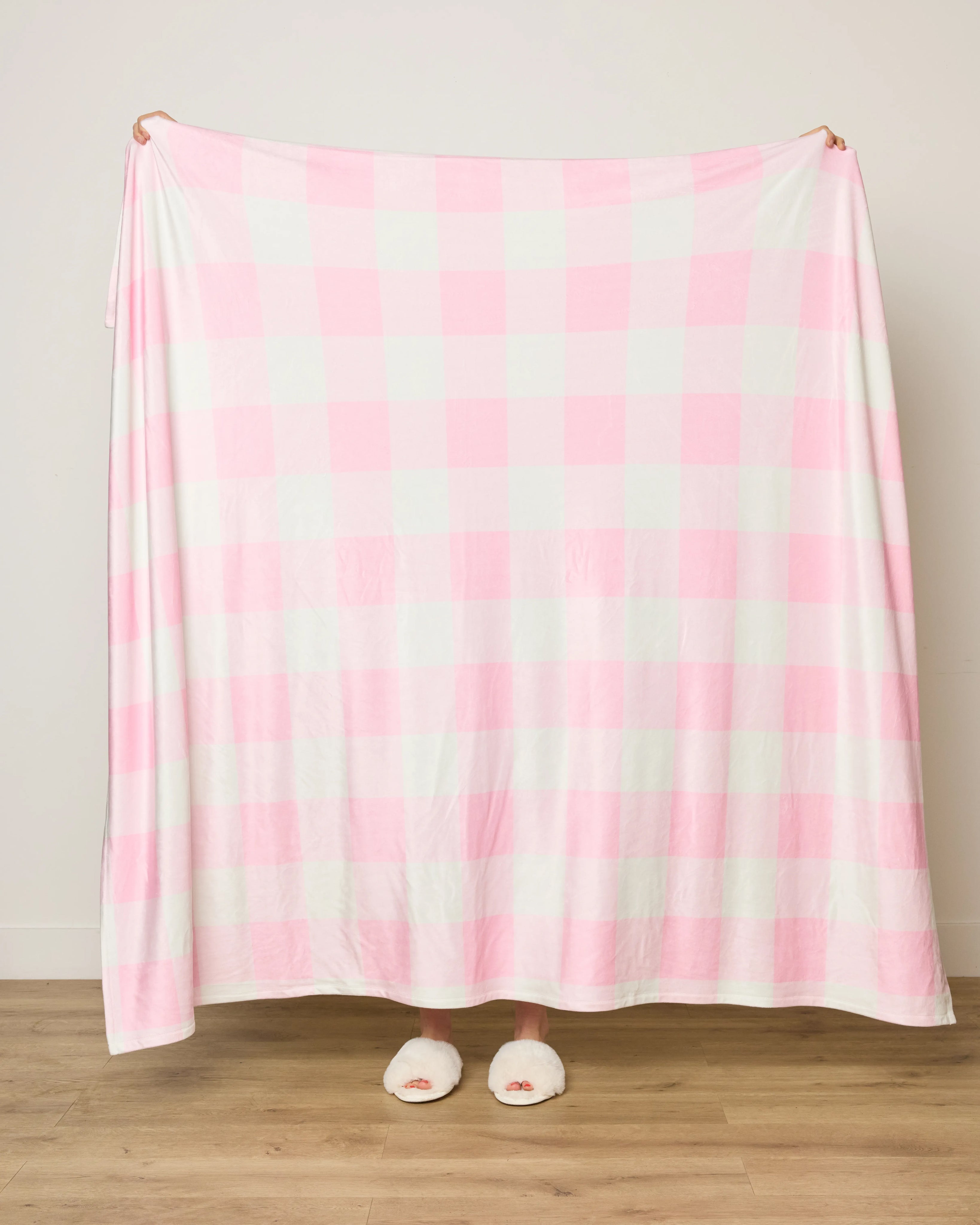 A person stands behind the Geometry Val Butter Blanket, large with a pink and white checkered pattern, hiding their body; only their hands and feet in white slippers are visible against a wooden floor.