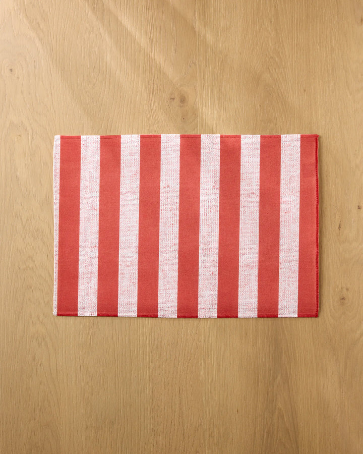 The Kailee Wright Peppermint Stripe placemat, featuring vertical red and white stripes, lies flat on a light wooden surface, adding a vibrant and eco-friendly touch to your dining space.