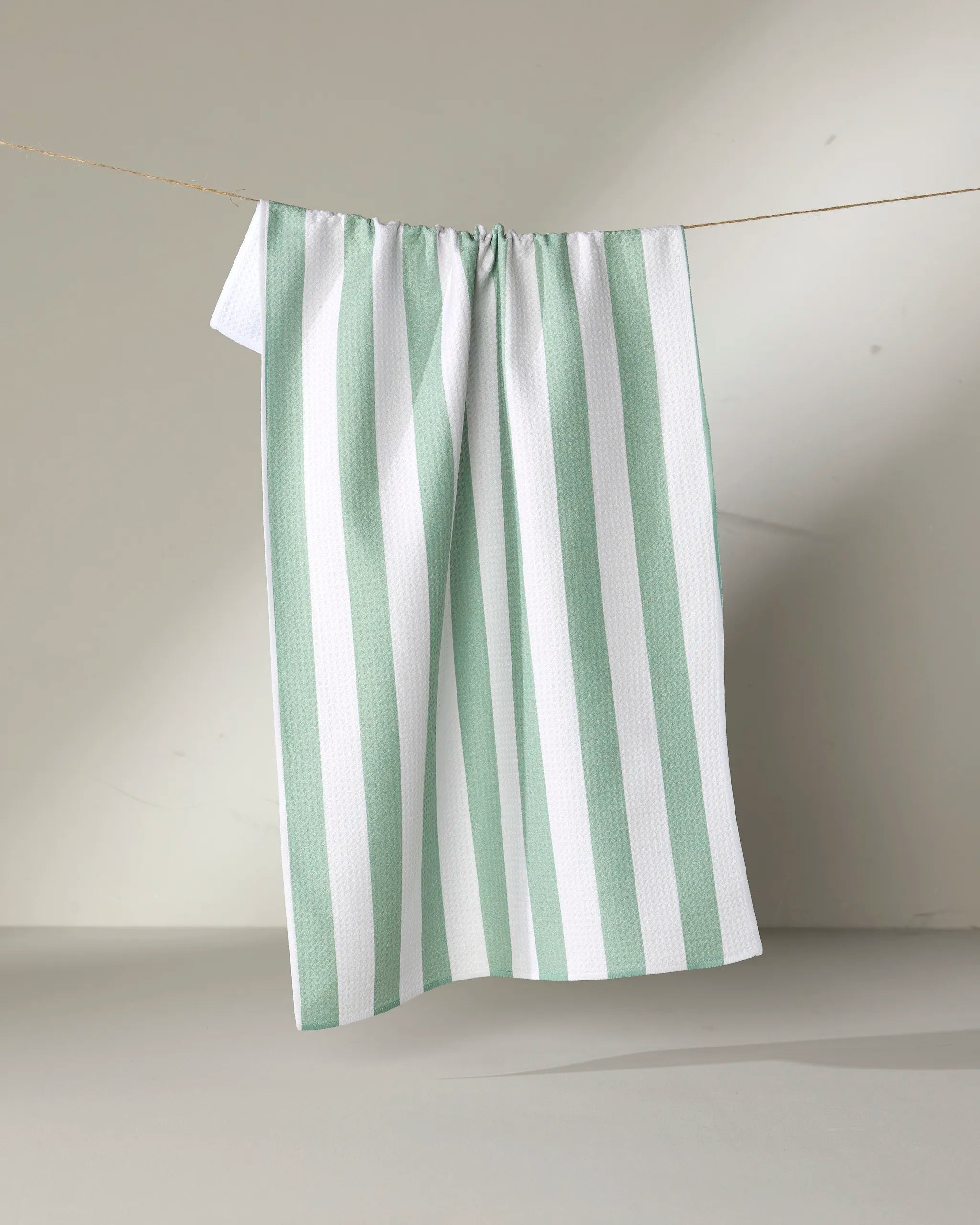 The Mistletoe Striped tea towel by Kailee Wright, made from recycled materials, features vertical light green stripes on white and hangs on a clothesline against a plain, light background with a soft shadow behind it.