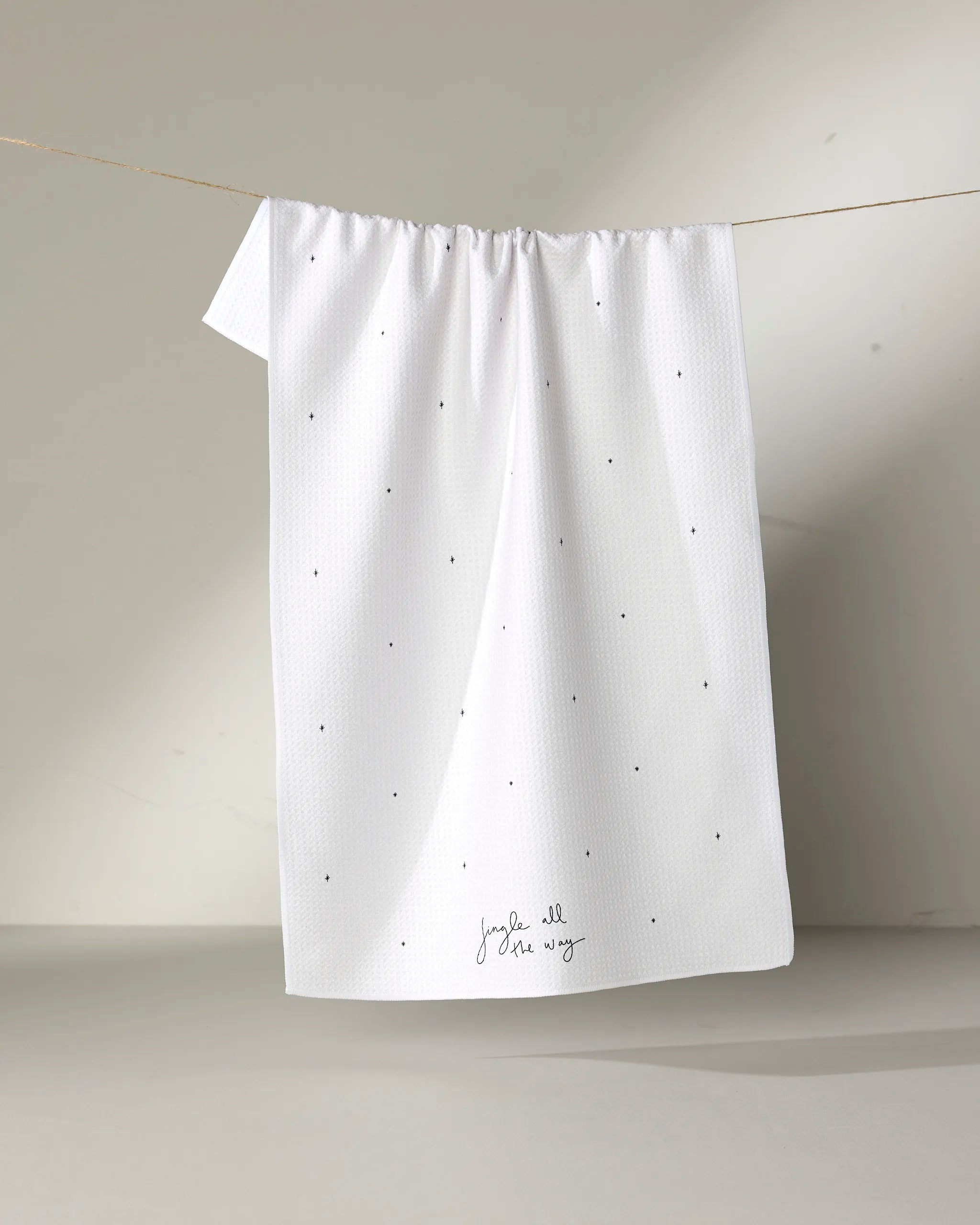 The Kailee Wright "Jingle All the Way" white kitchen towel, featuring small black dots and cursive text near the bottom, hangs on a thin string against a plain, light-colored background.