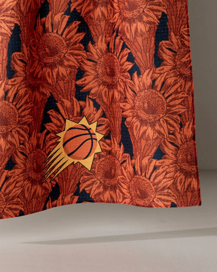 A close-up of the Suns Floral fabric by NBALAB x Geometry, featuring orange sunflowers and the Phoenix Suns logo on a dark background—ideal for an NBA-inspired tea towel.