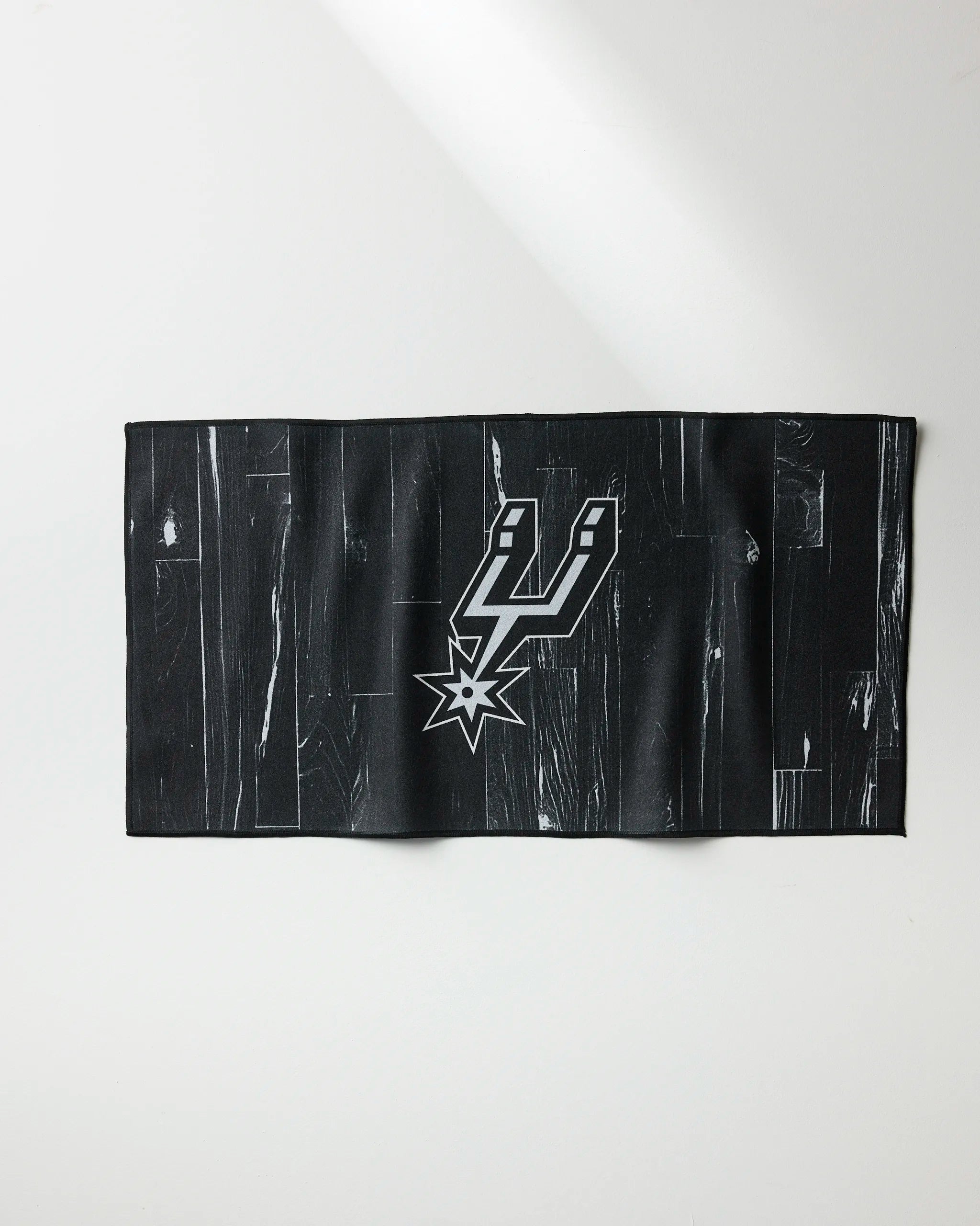 The Spurs Zone towel by NBALAB x Geometry is a black rectangular fitness cloth with a subtle wood grain pattern and a white and silver San Antonio Spurs logo at the center, perfect for fans and athletes.