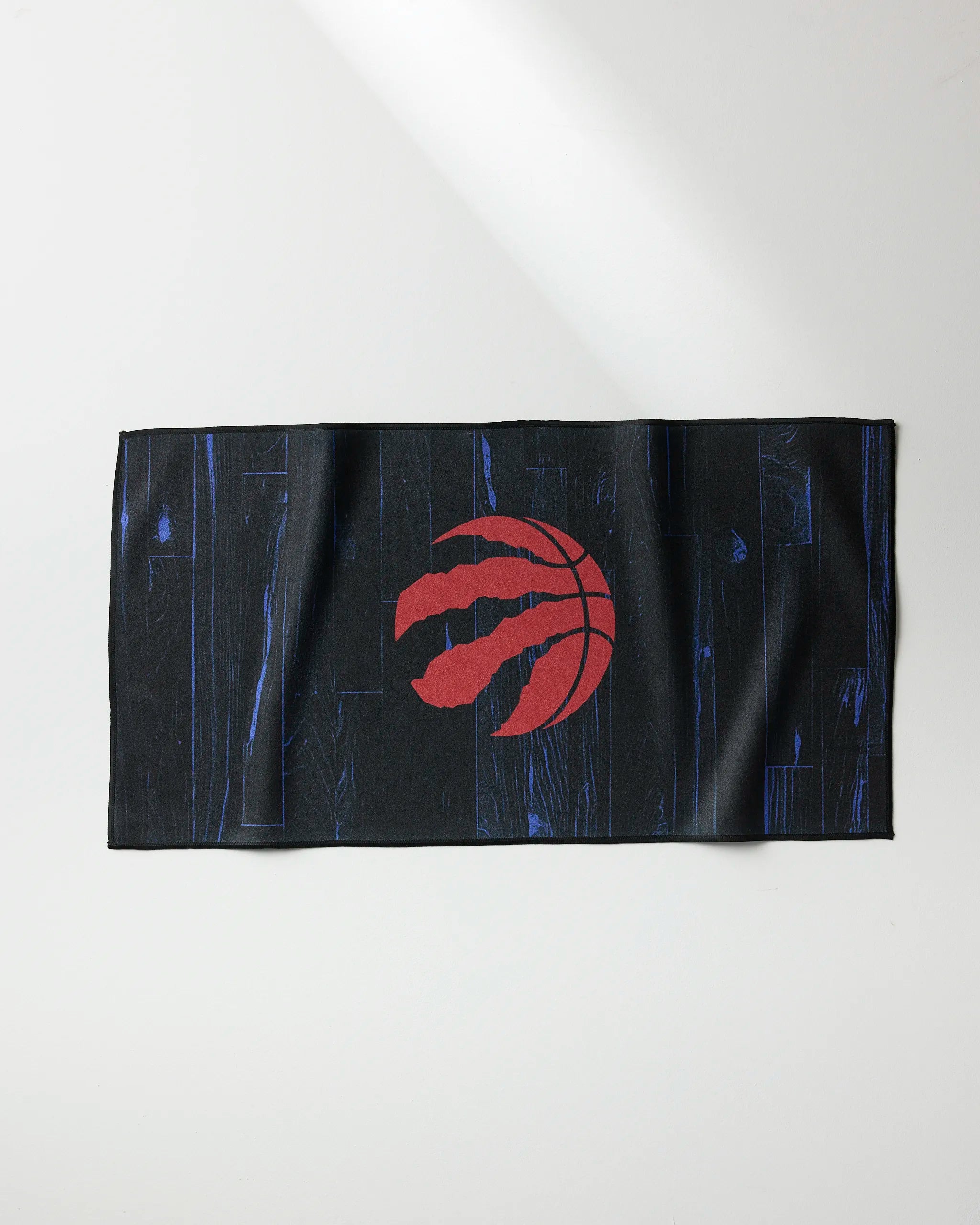 The Raptors Zone fitness towel by NBALAB x Geometry features a black design with a blue wood grain pattern and a red Toronto Raptors claw logo at the center, shown laid flat on a light surface.