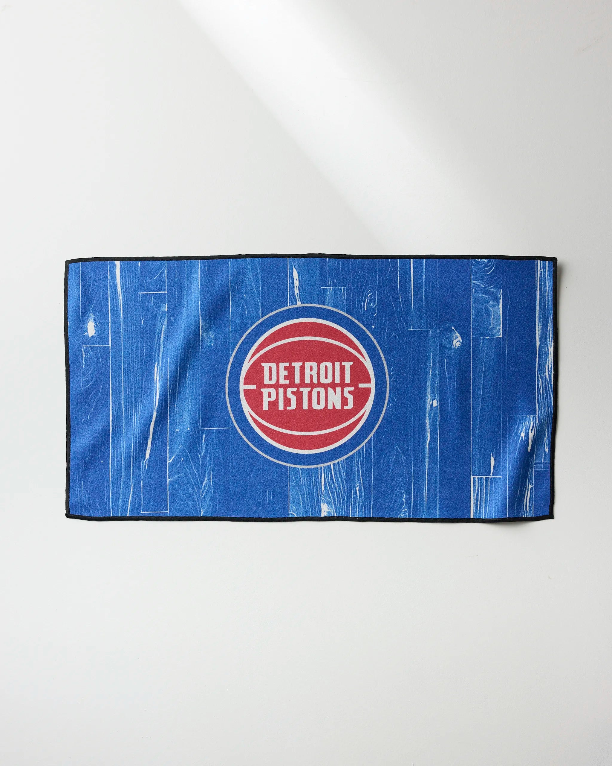 The Pistons Zone gym towel by NBALAB x Geometry features a blue wood plank design with the Detroit Pistons logo—a red circle with white text—centered on it. The rectangular towel is shown on a plain light background.