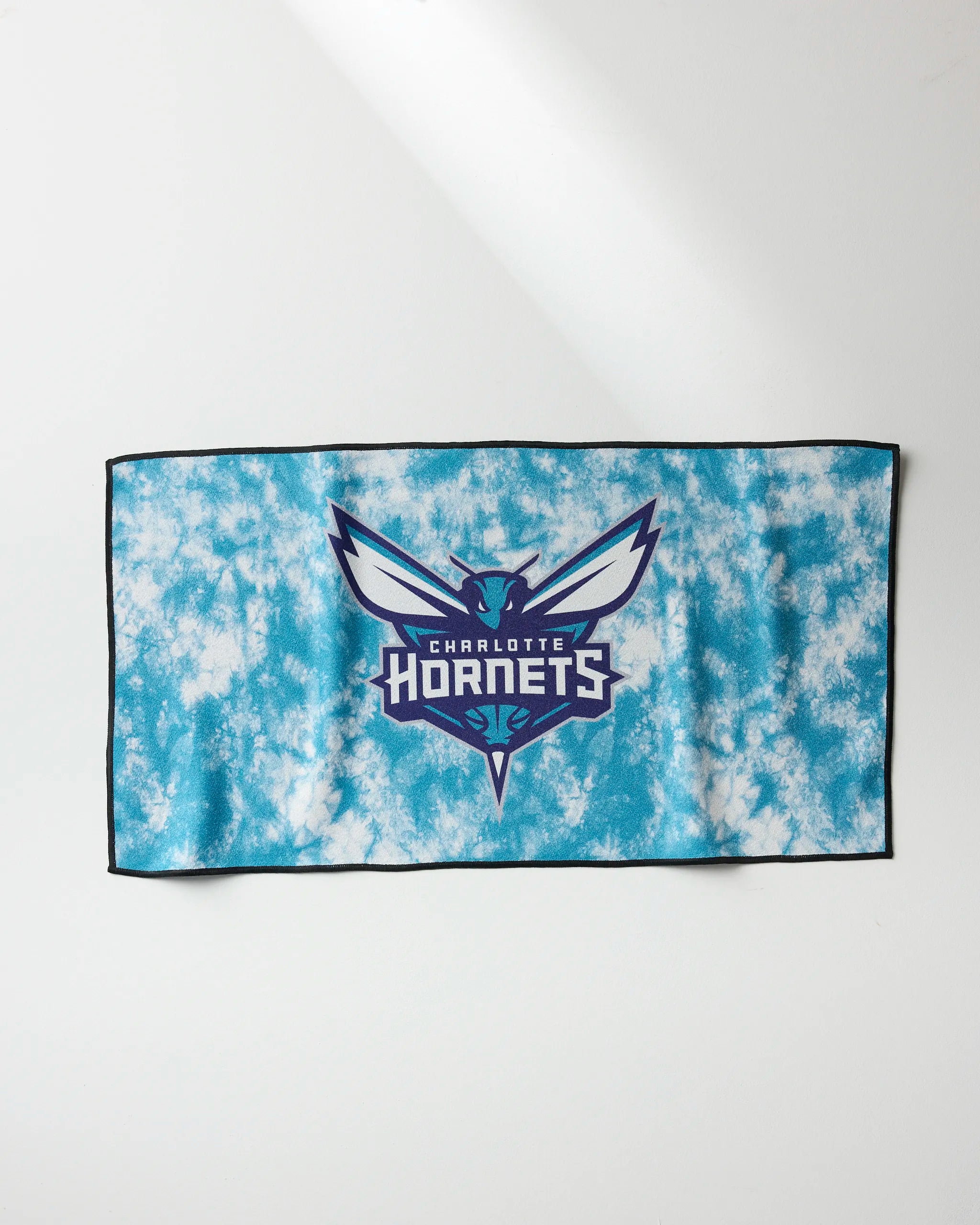 The Hornets Flash towel by NBALAB x Geometry is a rectangular, quick-drying gym towel with a blue and white tie-dye design, featuring the Charlotte Hornets logo and name centered, plus a black border. It’s shown flat on a white surface.