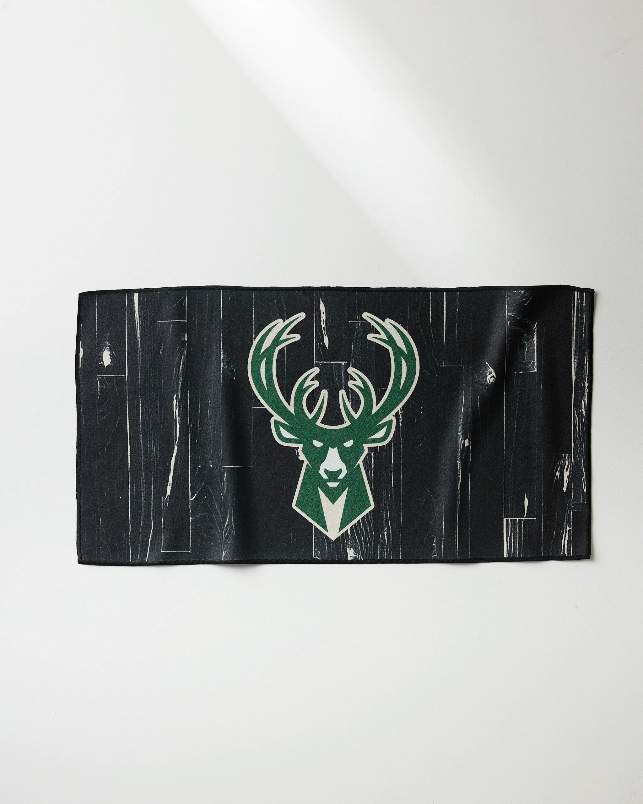 The NBALAB x Geometry "Bucks Zone" fitness towel features a black cloth with a distressed wood pattern and the Milwaukee Bucks green and white deer logo at the center, shown on a white surface.