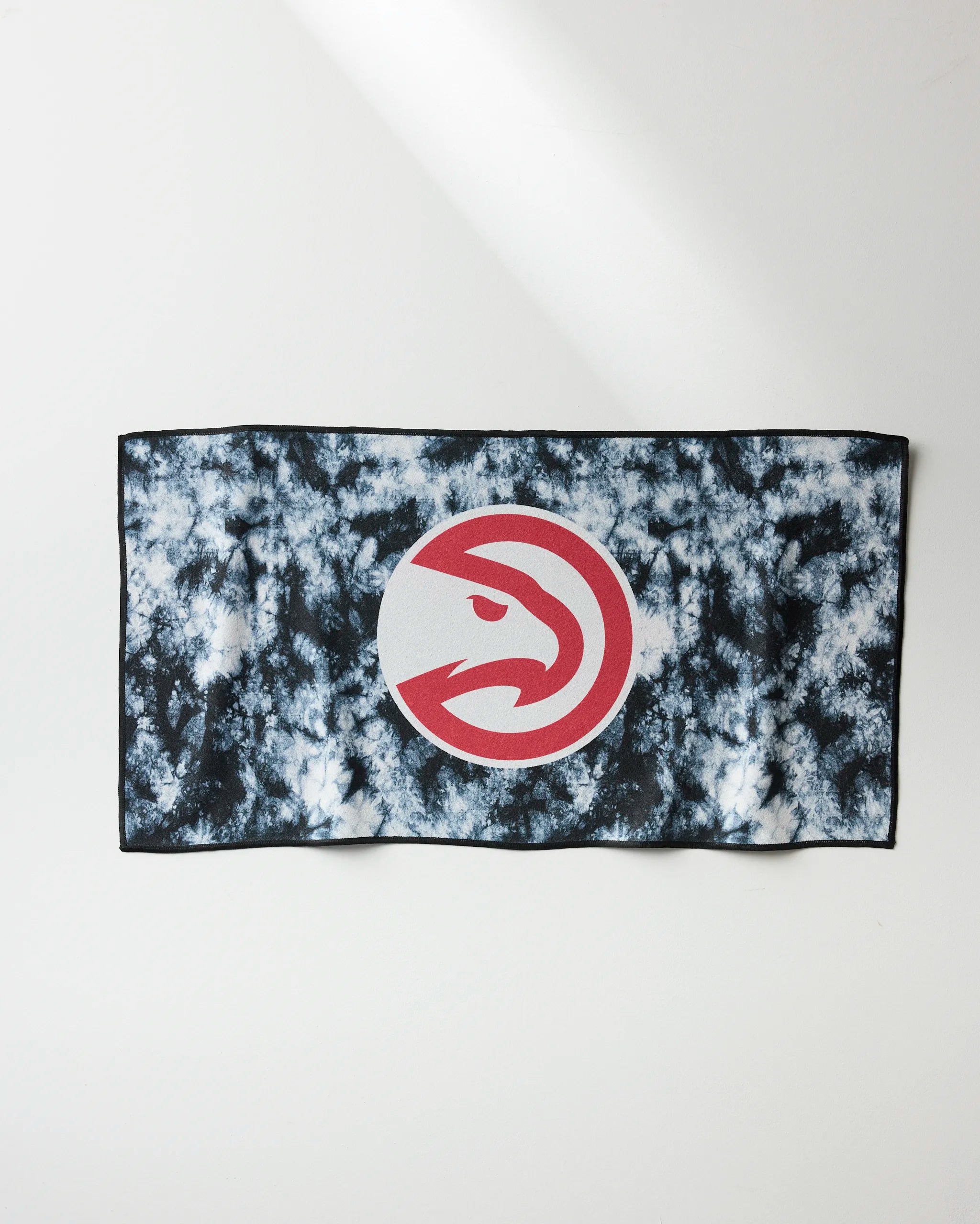 The NBALAB x Geometry "Hawks Flash" towel is a rectangular, quick-drying fitness towel with a black and white tie-dye pattern and a large red Atlanta Hawks logo at the center. It’s shown laid flat on a white surface.