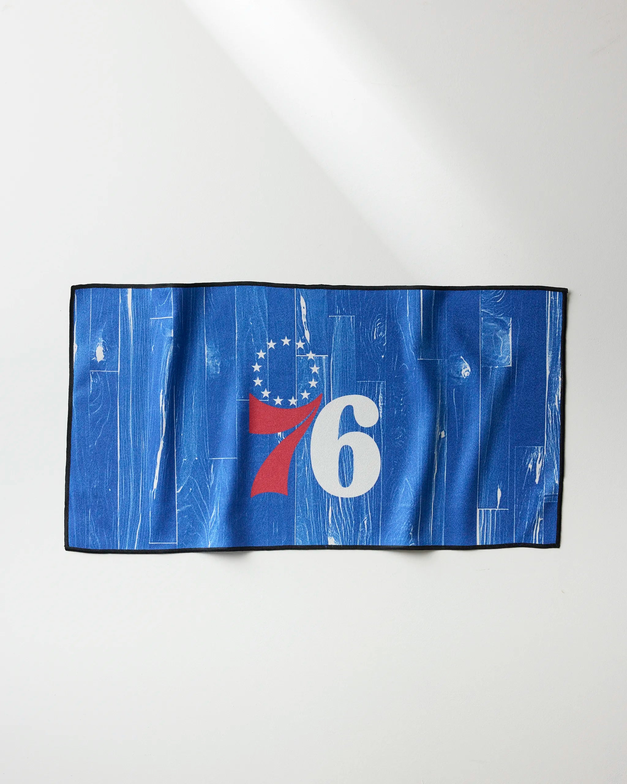 The NBALAB x Geometry 76ers Zone fitness towel, blue with a wood pattern, the number 76 in white and red, and thirteen stars above the 7 representing the Philadelphia 76ers, is displayed on a light surface.