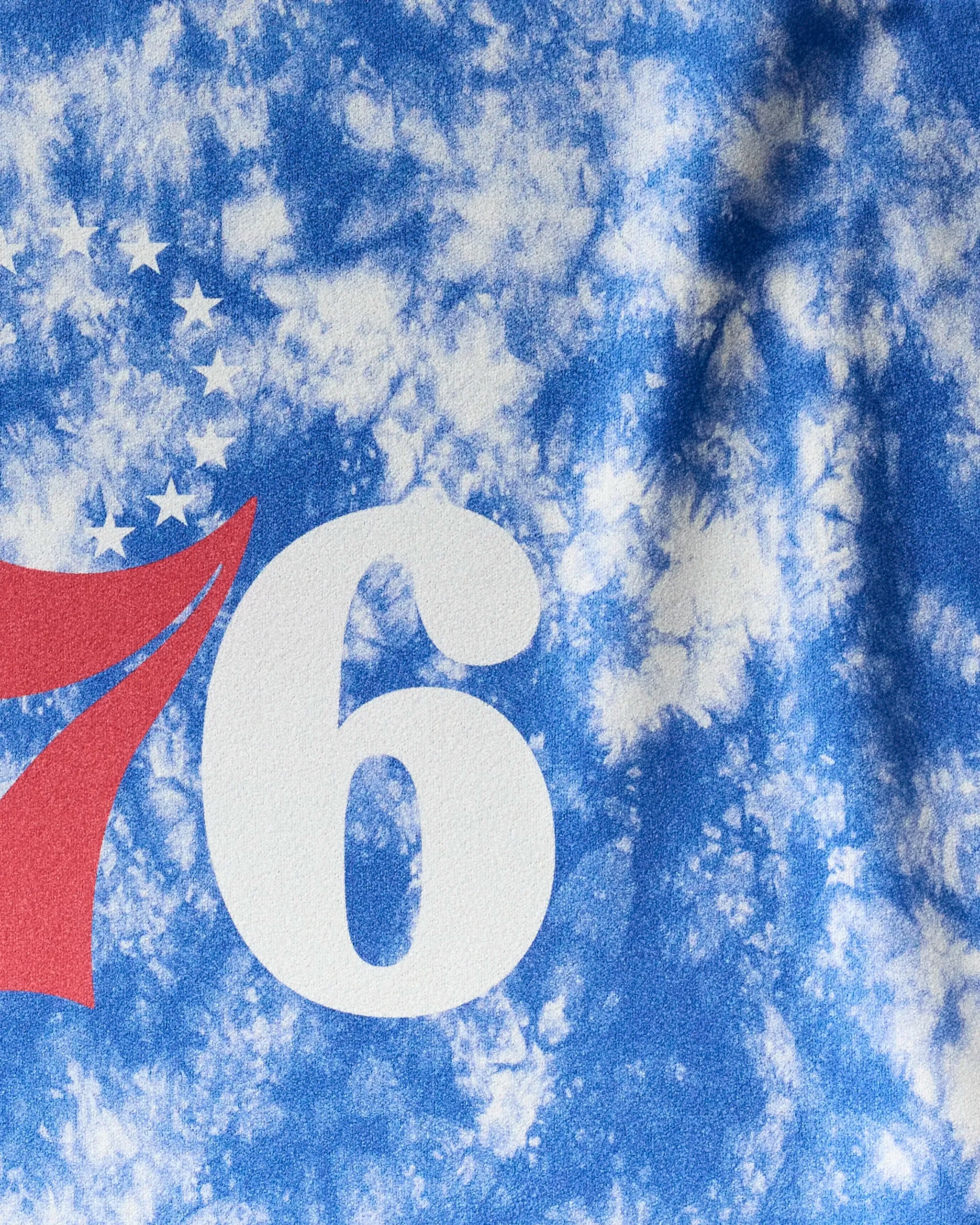 A close-up of the 76ers Flash fitness towel by NBALAB x Geometry features blue and white tie-dye, a partial red 7, a bold white 6, and a curved line of white stars.