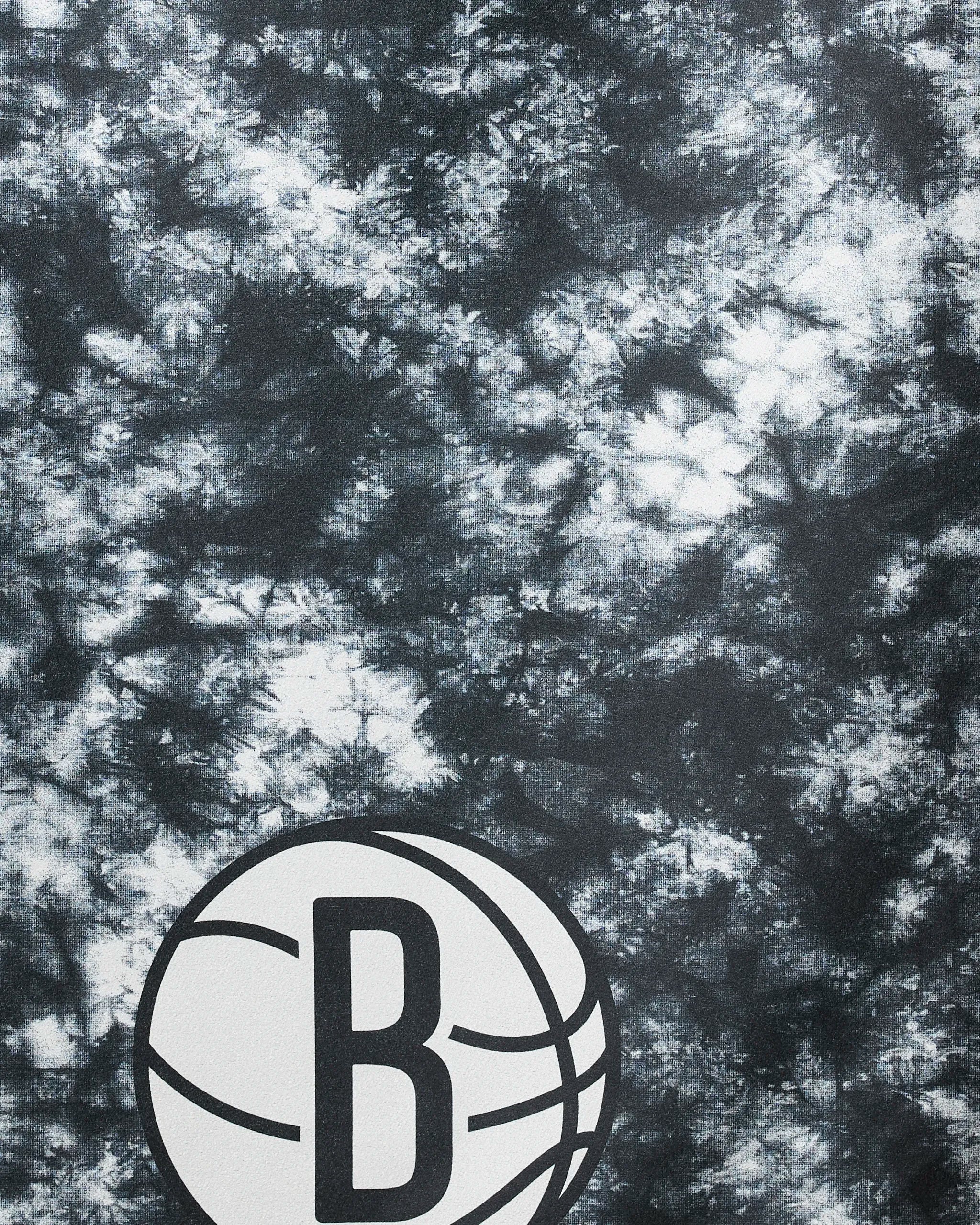 Close-up of the Nets Flash beach towel by NBALAB x Geometry, in black and white tie-dye, with a stylized B inside a basketball in the lower left corner representing the Brooklyn Nets.
