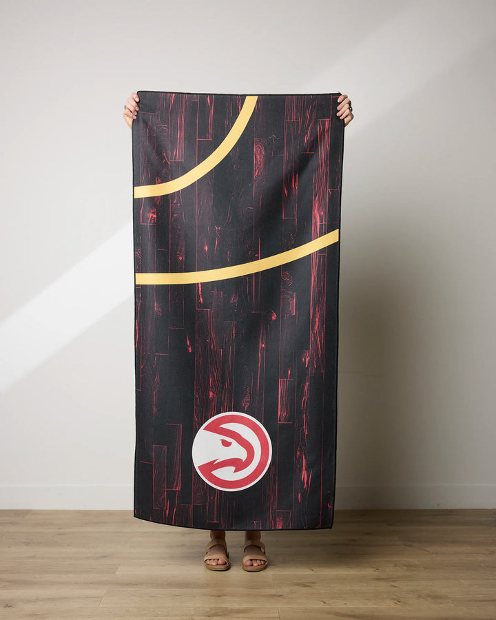 A person stands indoors holding the NBALAB x Geometry Hawks Zone towel, showing its black and red wood pattern, yellow curves, and Atlanta Hawks logo. Made from recycled materials; only their hands, feet, and sandals are visible.