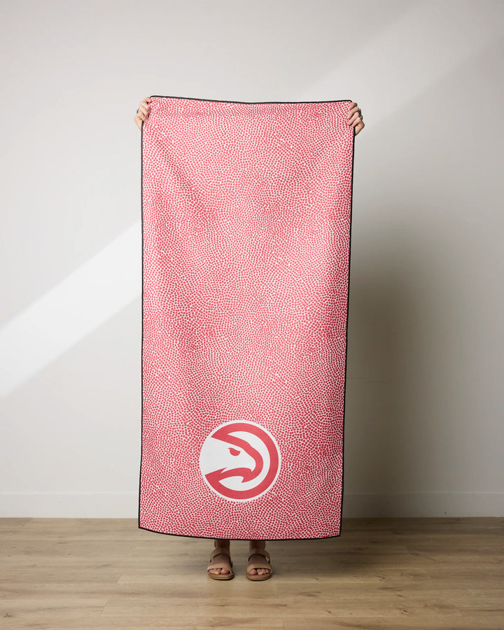 Indoors, a person hides their face and body behind the NBALAB x Geometry Hawks Grip quick-drying NBA beach towel featuring a red and white dotted pattern with the Atlanta Hawks logo near the bottom.
